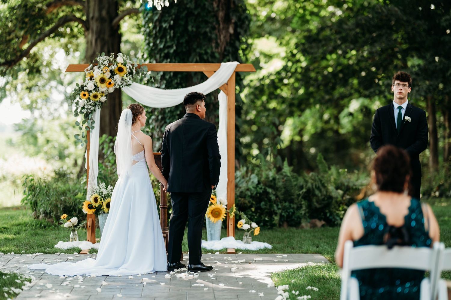 Outdoor wedding ceremony with couple at sunflower-adorned wooden arch, officiant, and guest in teal dress watching.