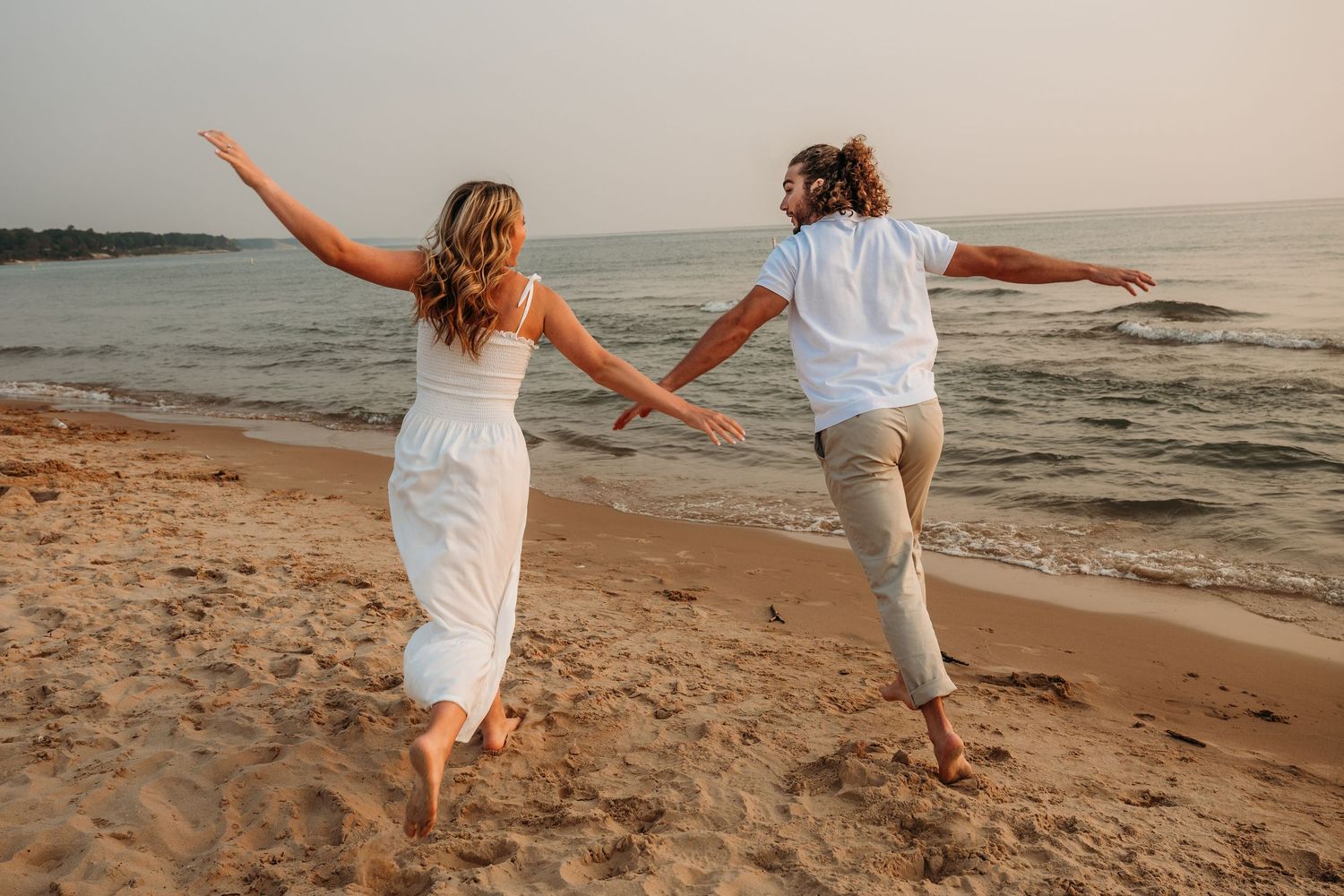 Two women joyfully running barefoot on a sandy beach at sunset, arms outstretched, wearing white and light casual outfits.