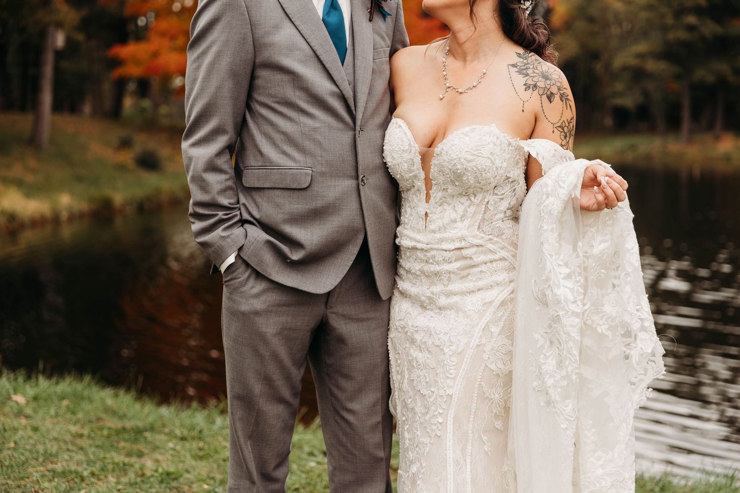 Couple in formal wedding attire stands together outdoors with autumn foliage and a lake in the background.