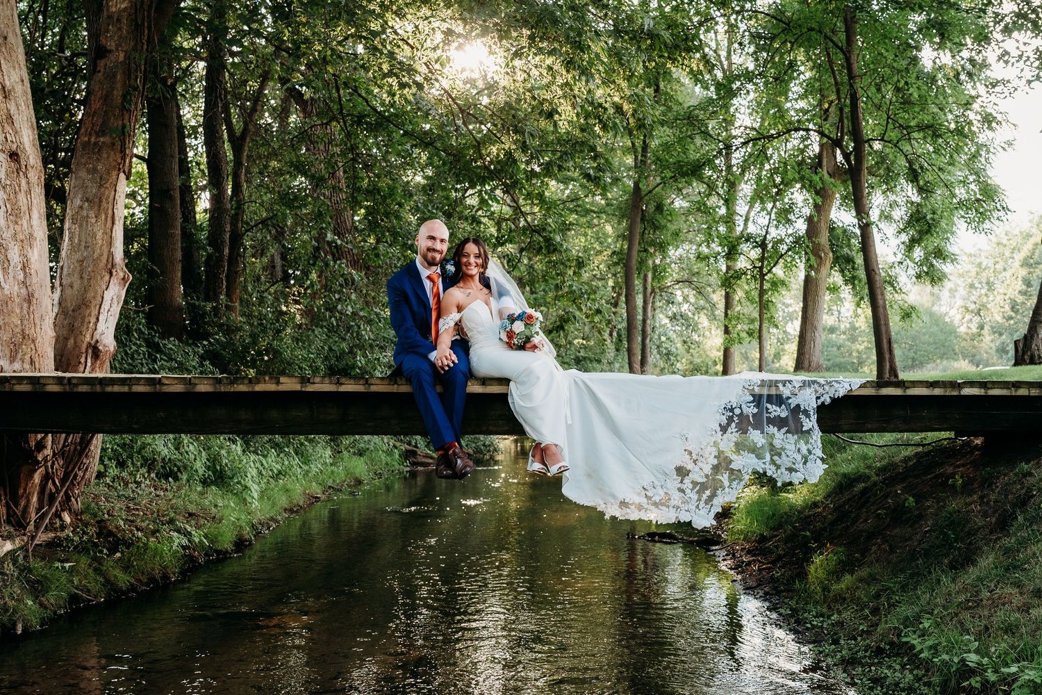 A couple in wedding attire poses together in a lush green forest setting during their outdoor ceremony.