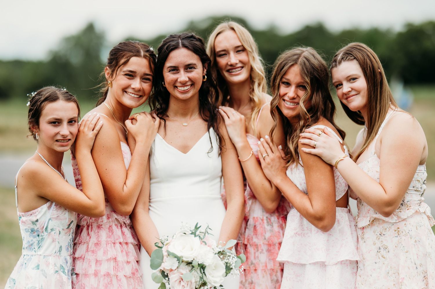 A group of bridesmaids in pink and white dresses smile and laugh together during an outdoor wedding photoshoot.