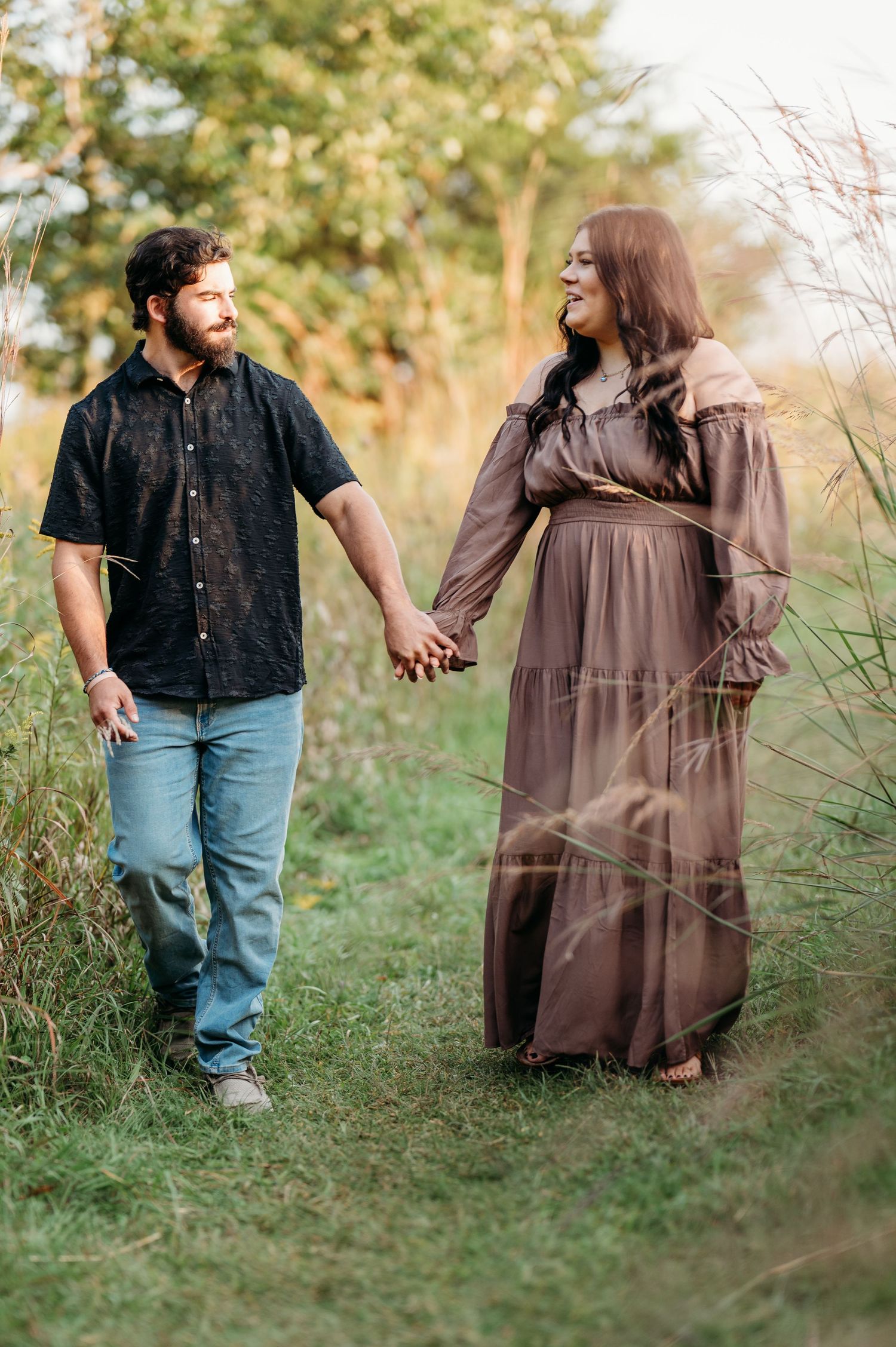 A couple holds hands while walking through a grassy meadow on a sunny afternoon, captured in a romantic photo series.
