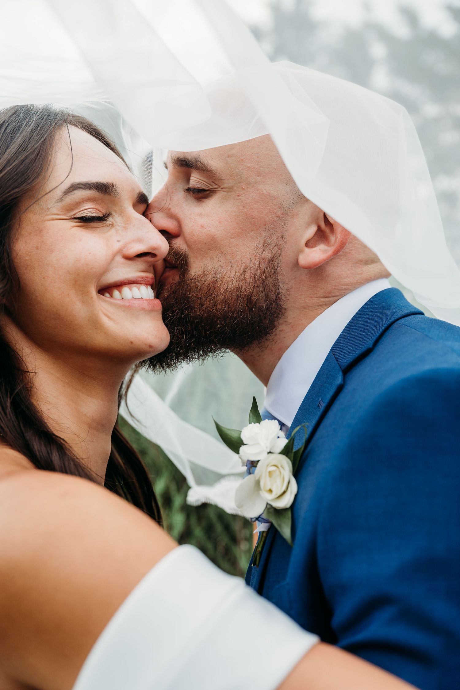 Groom in blue suit kissing bride on cheek under wedding veil, bride smiling with eyes closed.