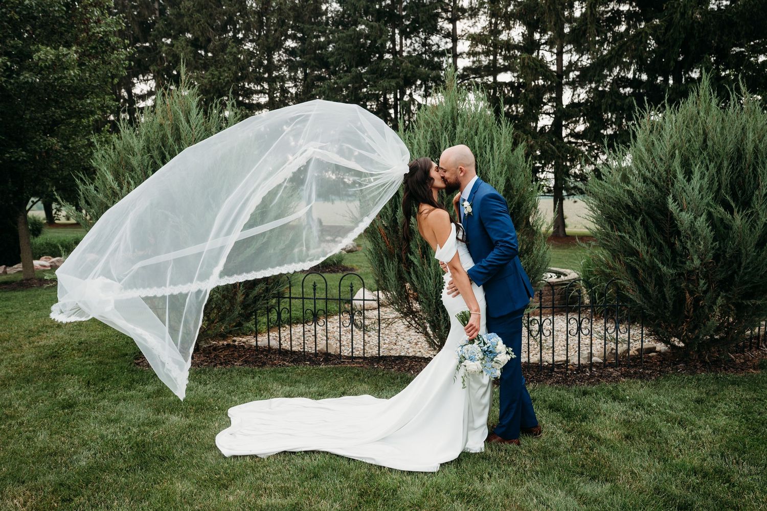 Bride and groom kissing outdoors as wedding veil billows in wind, lush green trees and iron fence backdrop.