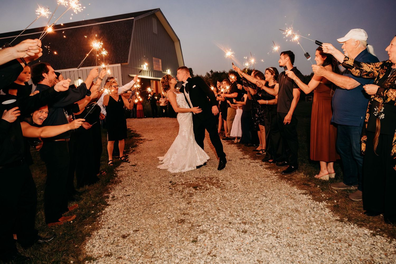 Bride and groom walk through sparkler tunnel surrounded by wedding guests at night outdoor barn venue celebration.
