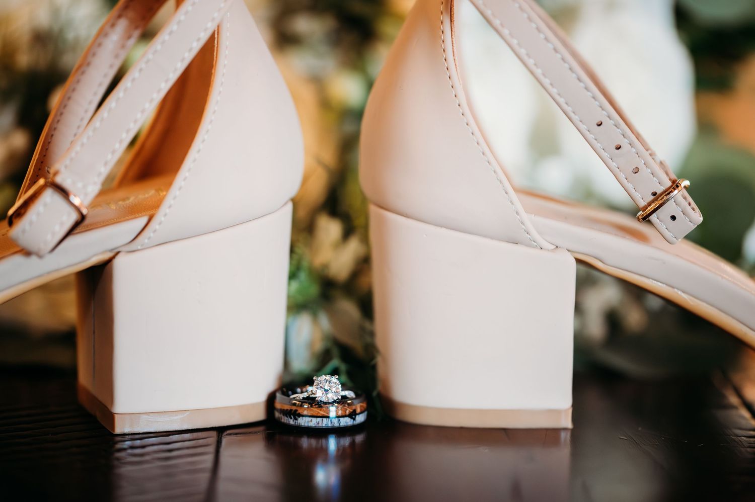 Wedding rings displayed between elegant ivory colored block heel shoes with ankle straps on dark wooden surface.
