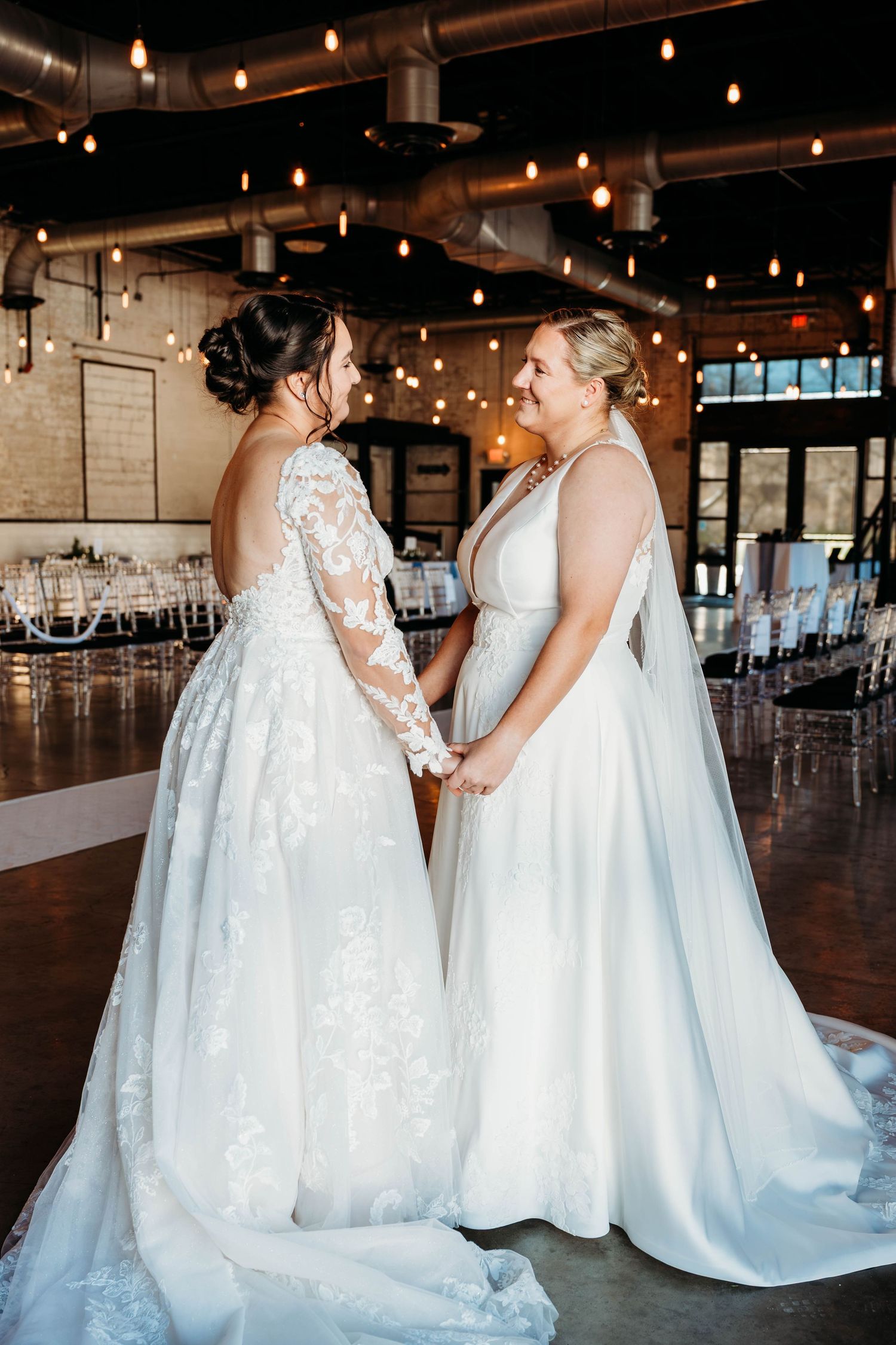 Wedding party in flowing white gowns standing in an industrial venue with string lights.