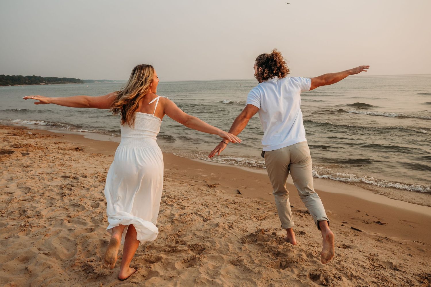 Carefree couple spreading their arms while walking on a sunset beach in white and beige outfits.