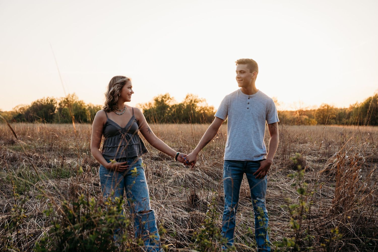 Two people holding hands stand together in a golden field at sunset.