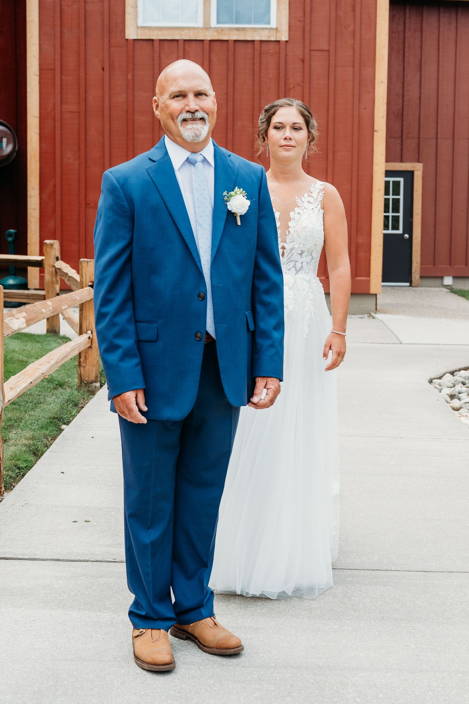 A couple poses outside a red barn on their wedding day, wearing a blue suit and white lace gown.