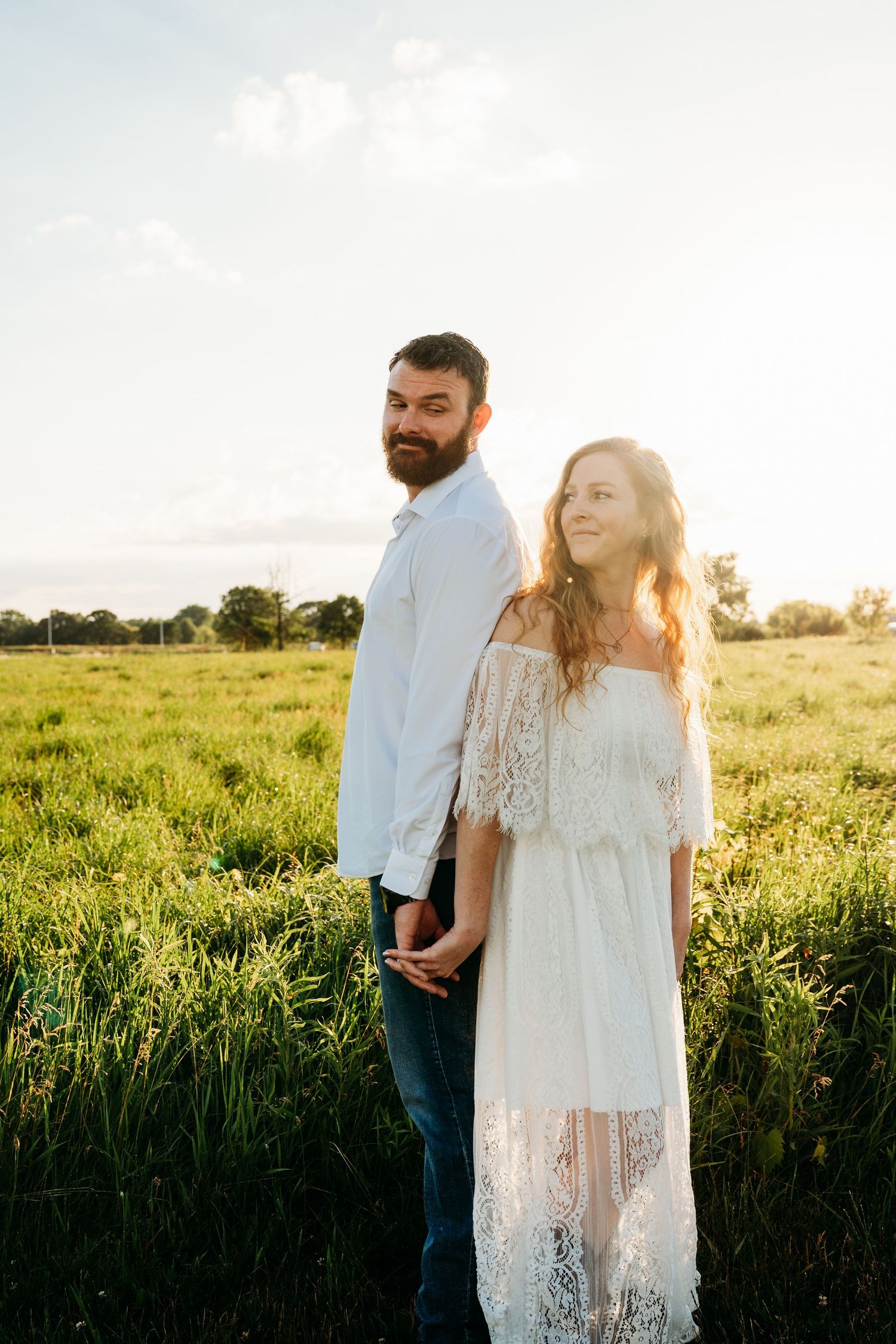 Couple holding hands in a sunlit green field, woman in white lace dress, man in white shirt, golden hour backdrop.