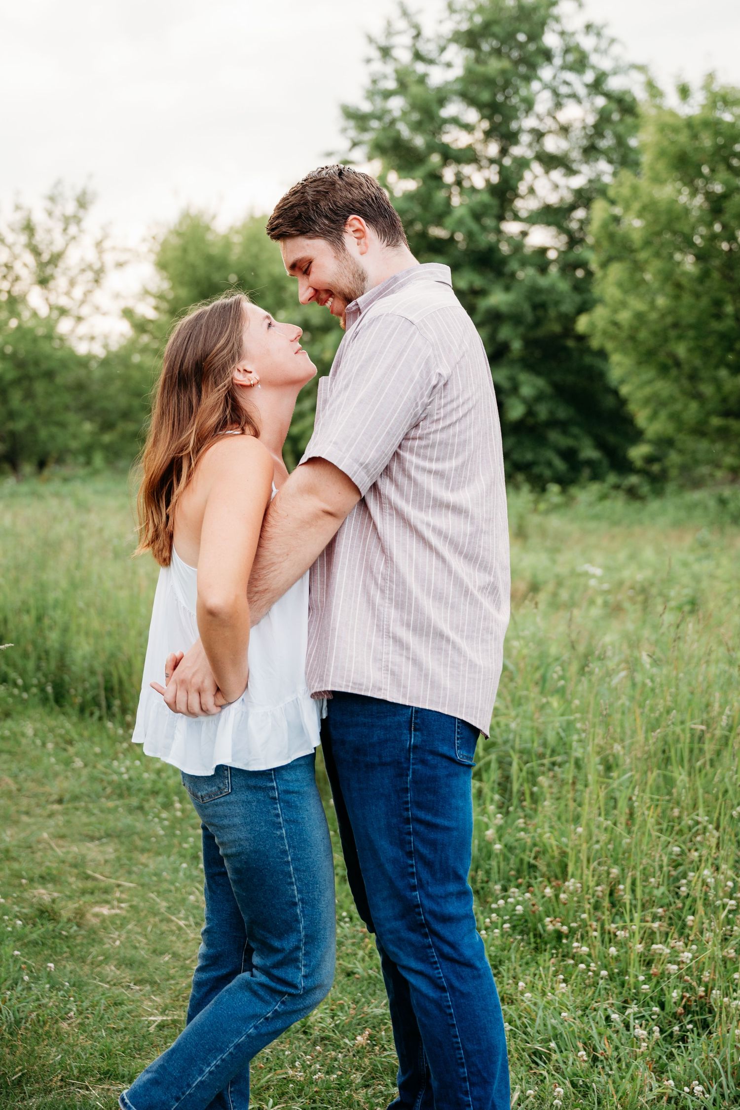 A couple smiles at each other while embracing in a lush green field, wearing casual jeans and light tops.