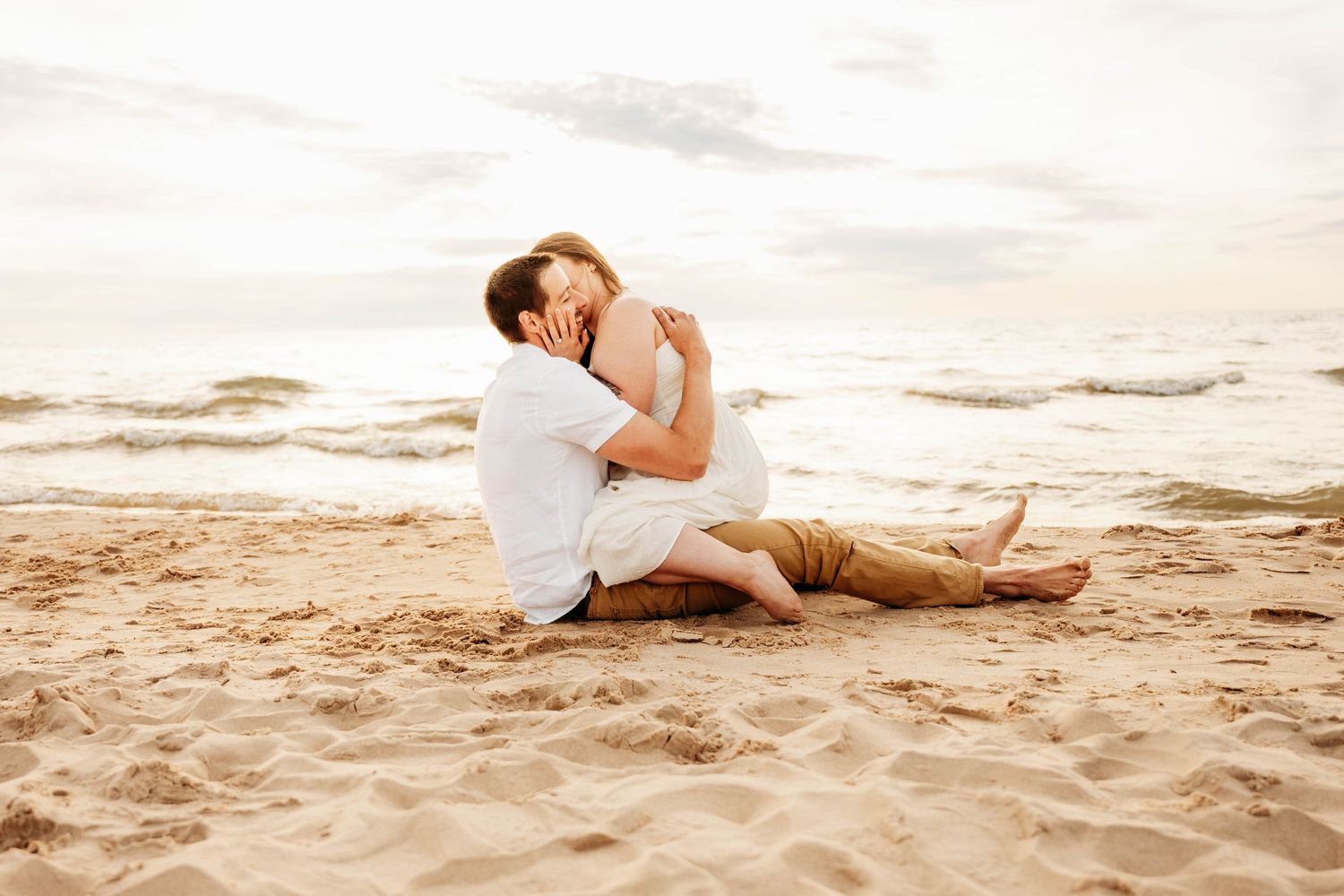 Romantic couple embracing on sandy beach at sunset, sitting together near ocean waves in casual white and tan outfits.