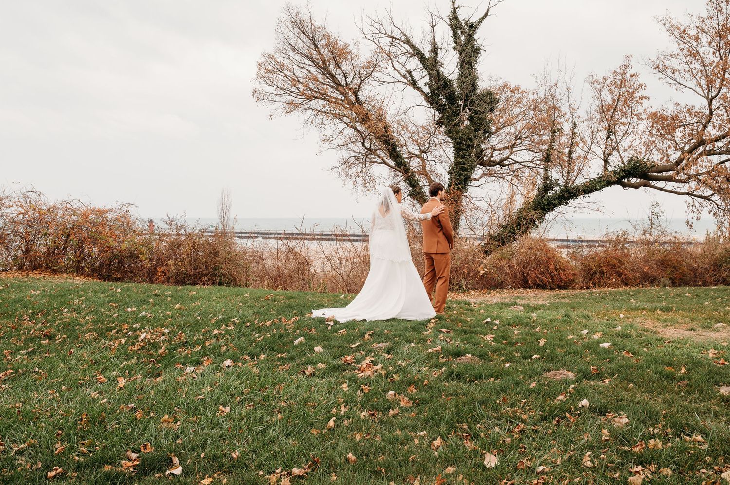 Bride and groom embrace near a rustic tree on a green lawn with autumn leaves during an outdoor fall wedding.