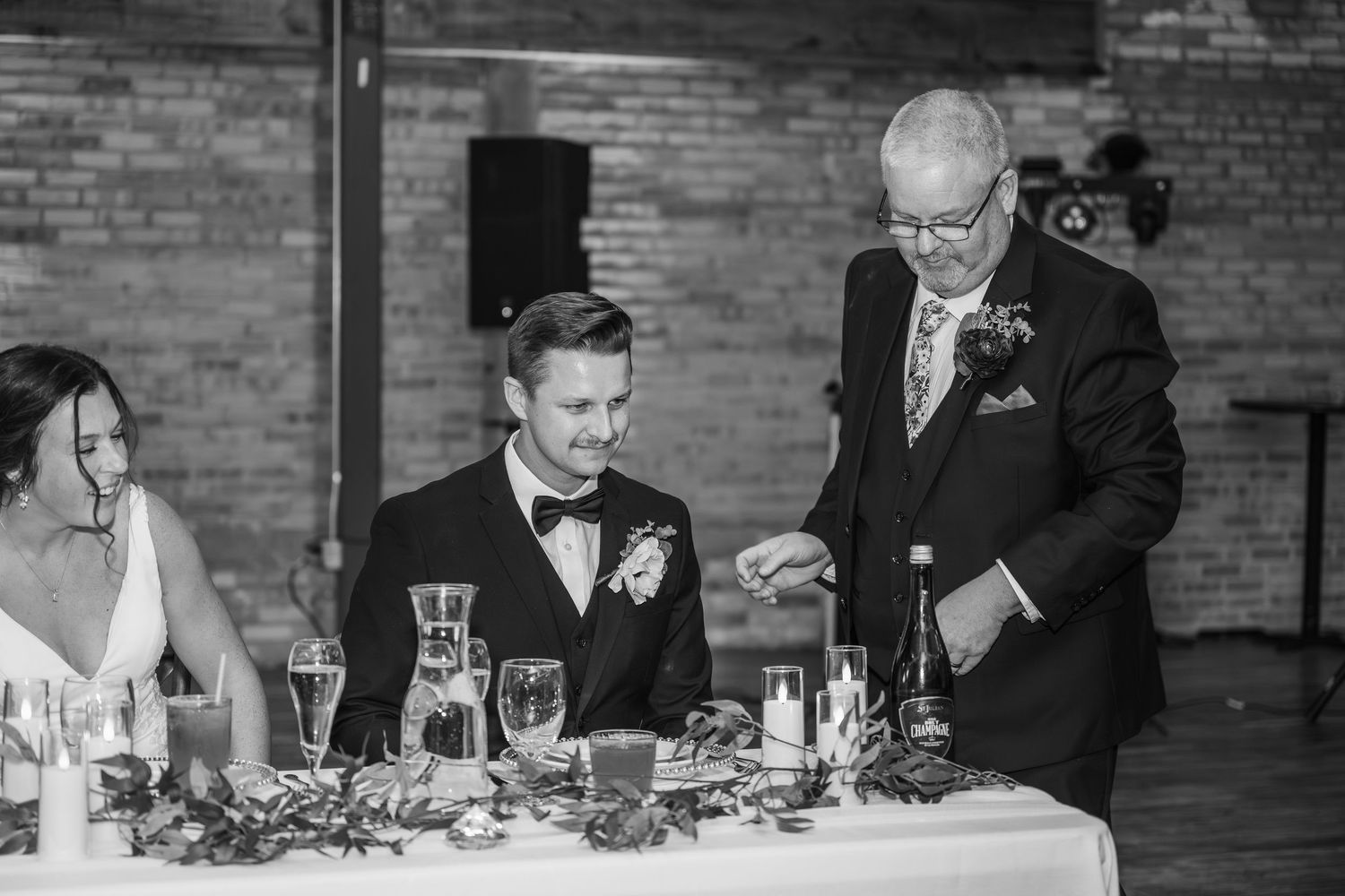 Black and white wedding reception photo of groom laughing with older man in suit at decorated head table.