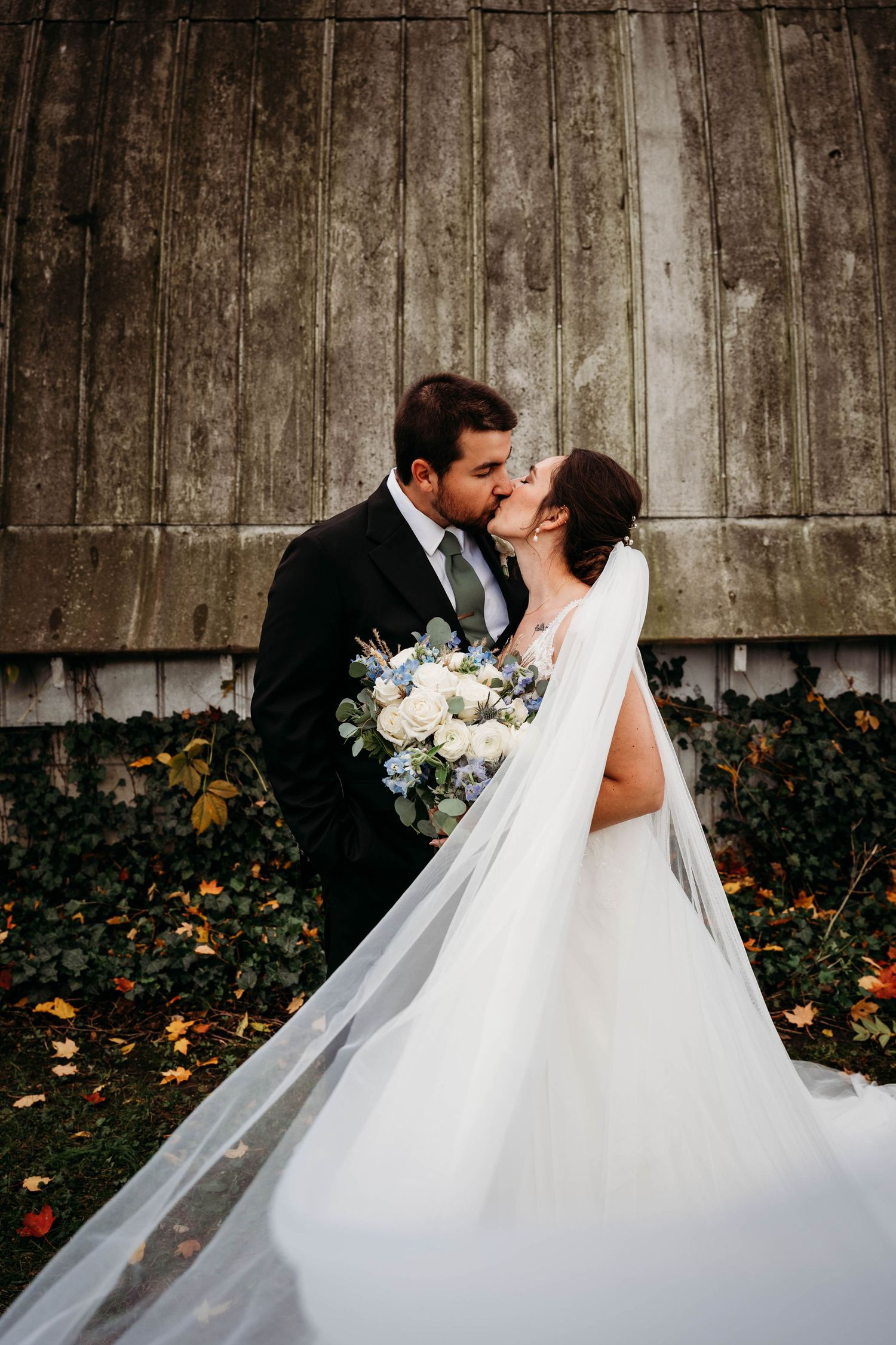 A romantic wedding couple embraces against a rustic wooden barn wall with flowing white veil and floral bouquet.