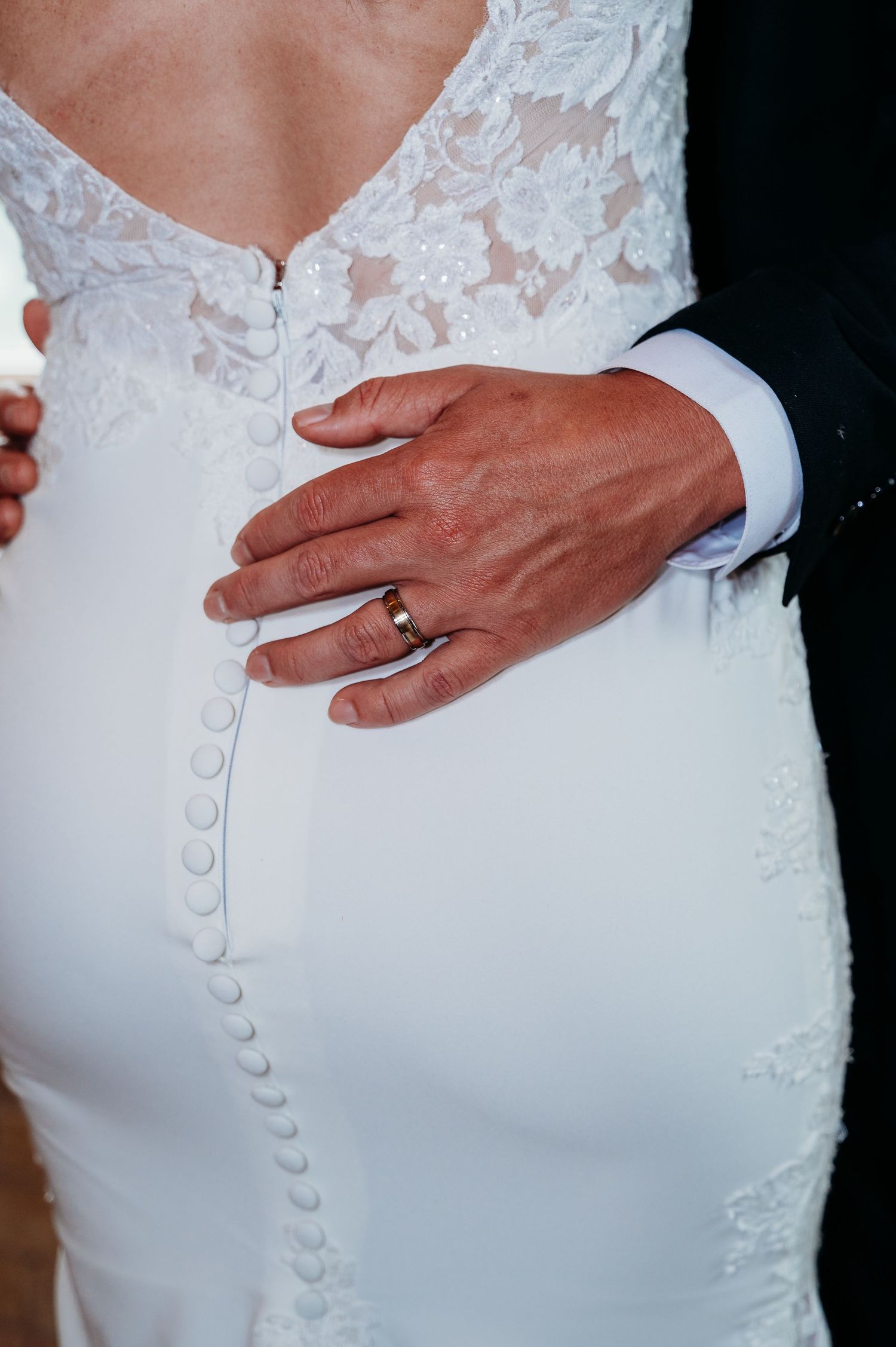 Close-up view of white wedding dress buttons and lace detail with hand wearing gold ring.