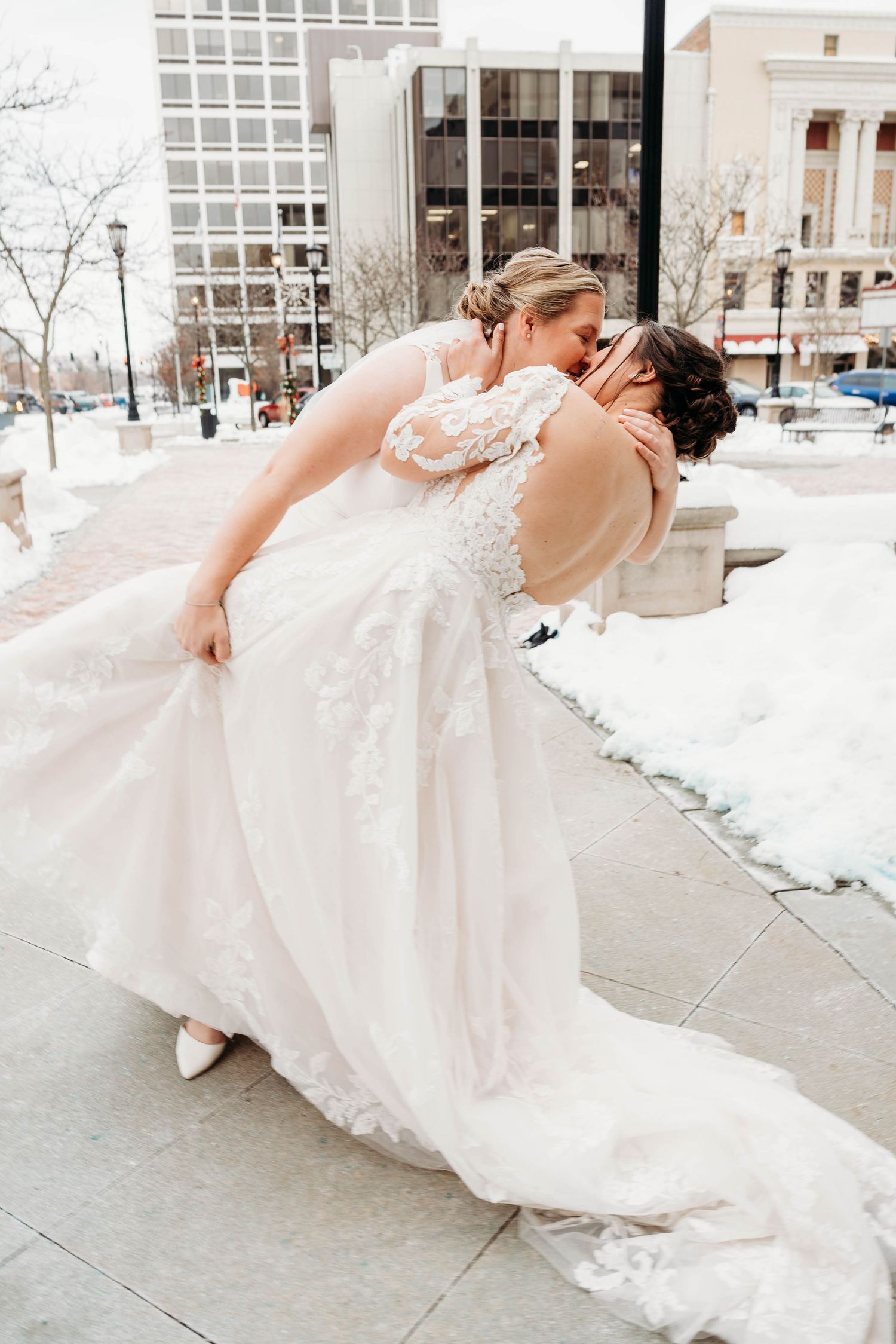 Two people in elegant white wedding dresses share a romantic kiss outdoors on a winter day with snow on the ground.