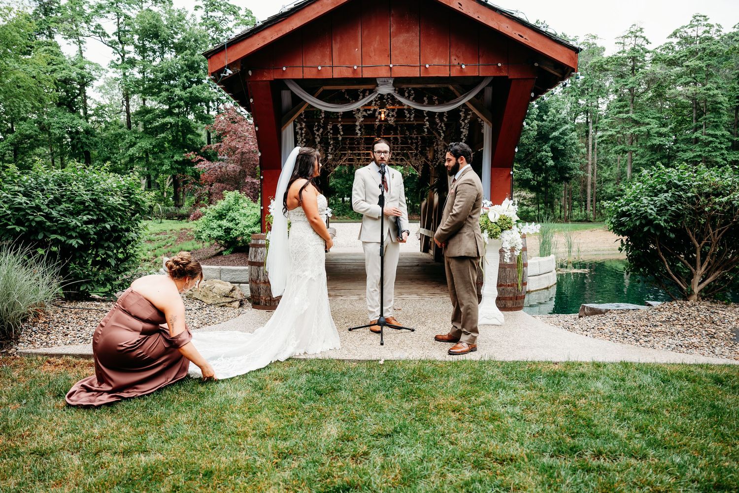 Outdoor wedding ceremony at a rustic red barn pavilion with bride, groom, and officiant on a lush green lawn.