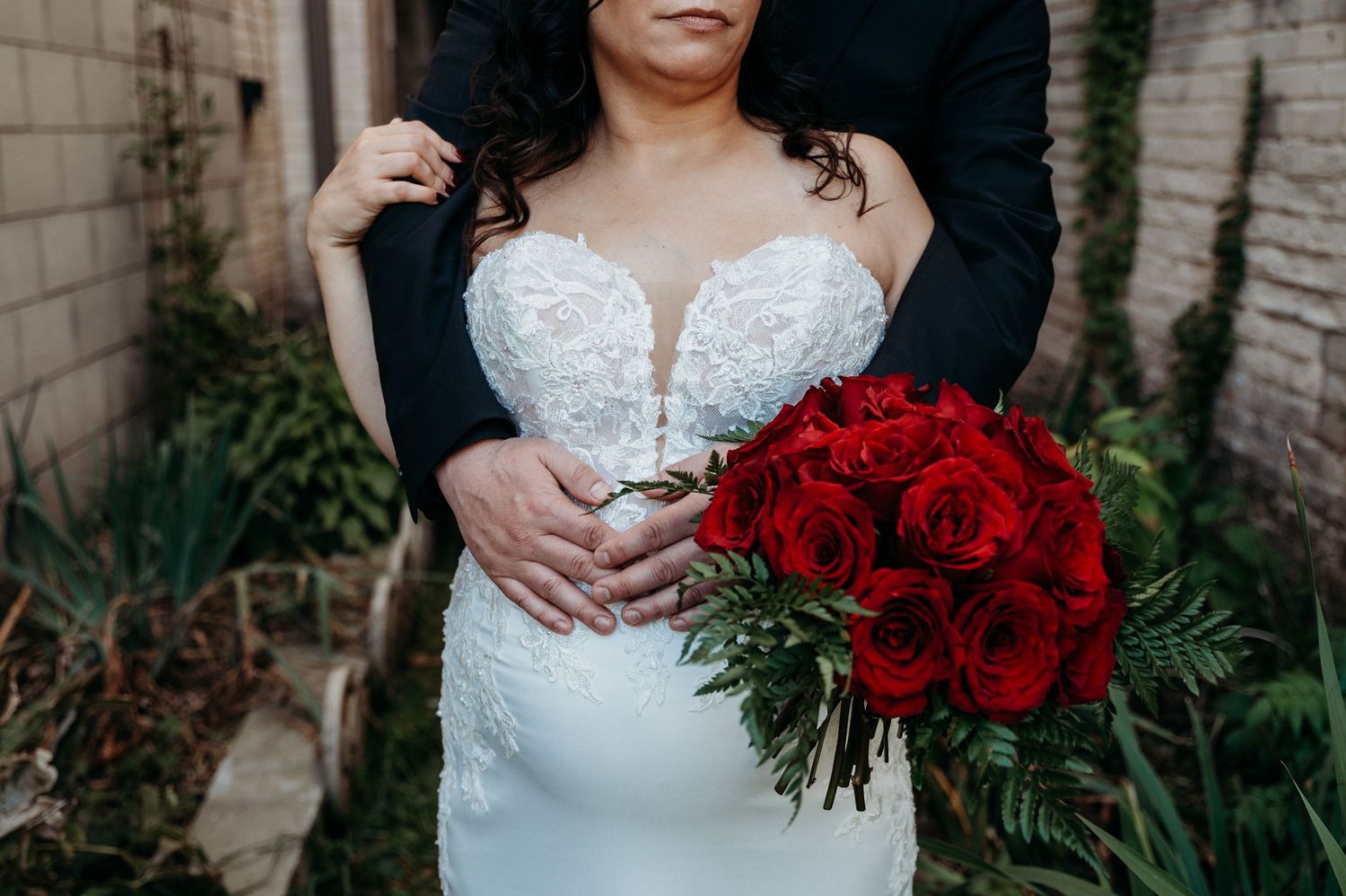 A couple in wedding attire poses with a vibrant bouquet of red roses as hands embrace in a romantic moment.