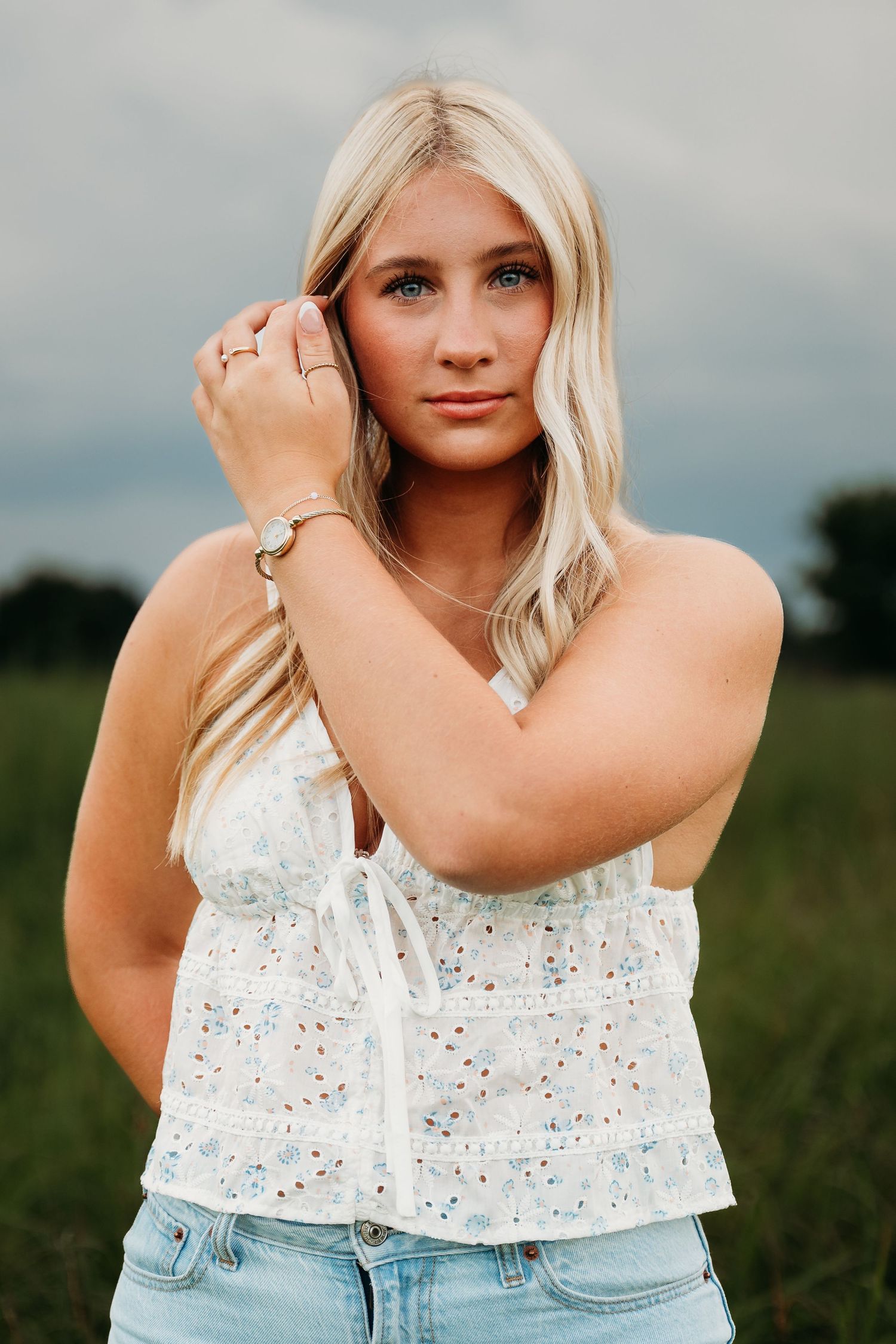 A person wearing a white floral top and denim shorts poses in a rural field setting.