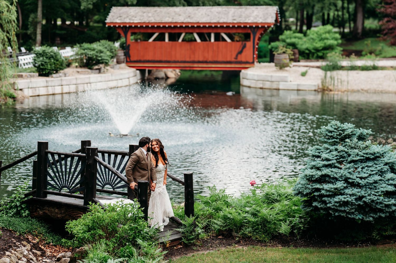 Couple sharing a romantic kiss on a garden bridge beside a serene pond with fountain and red covered bridge backdrop.