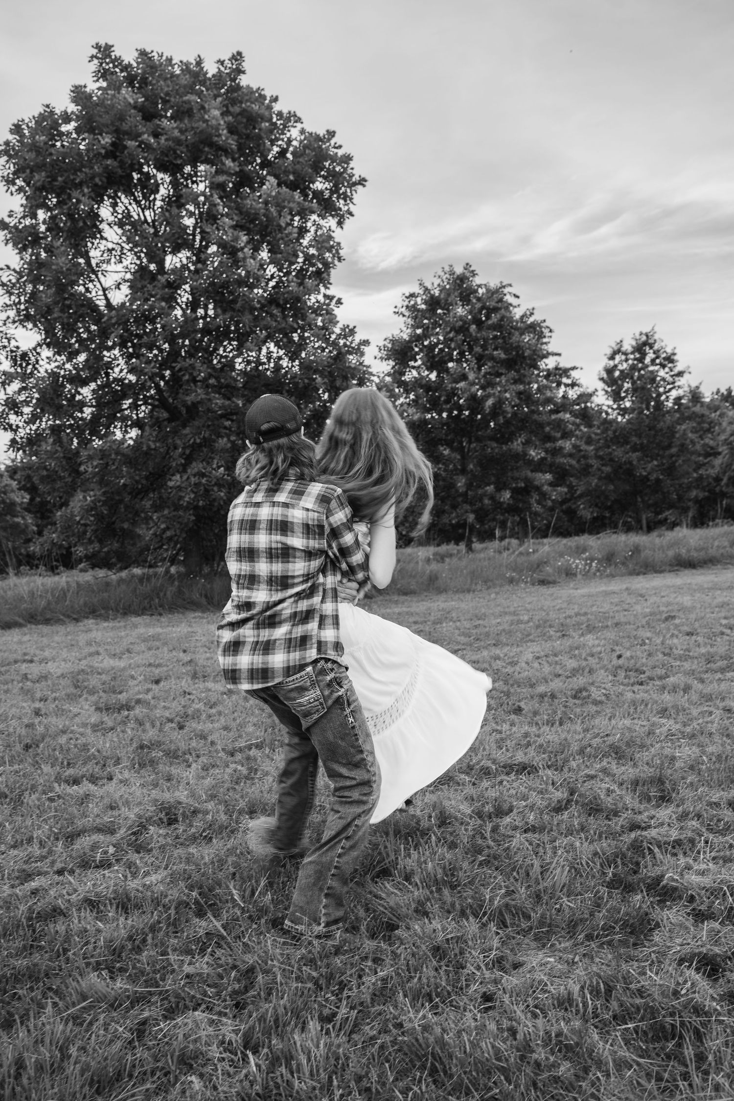 Black and white photo of a couple dancing in a field, man lifting woman in a white dress near trees.