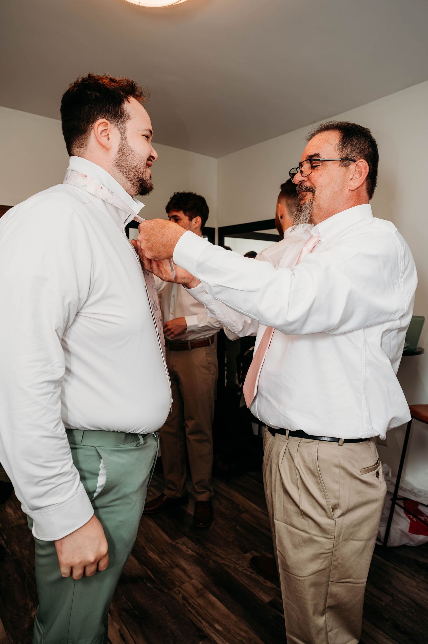 Man helping groom adjust bow tie while getting ready for wedding ceremony indoors.
