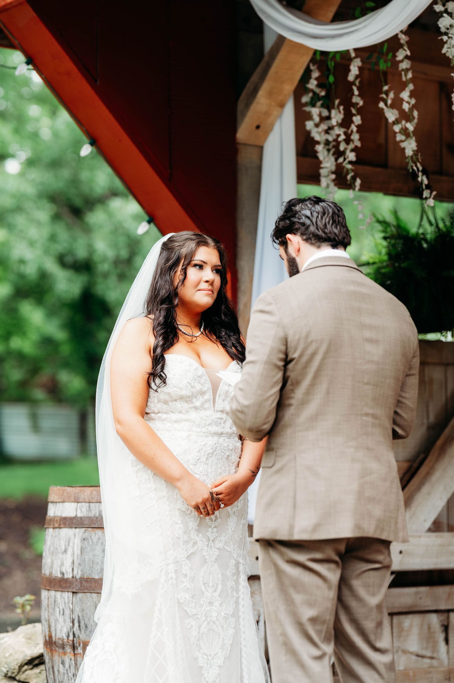 Bride in lace wedding gown and veil holds hands with groom in tan suit during outdoor first look ceremony.