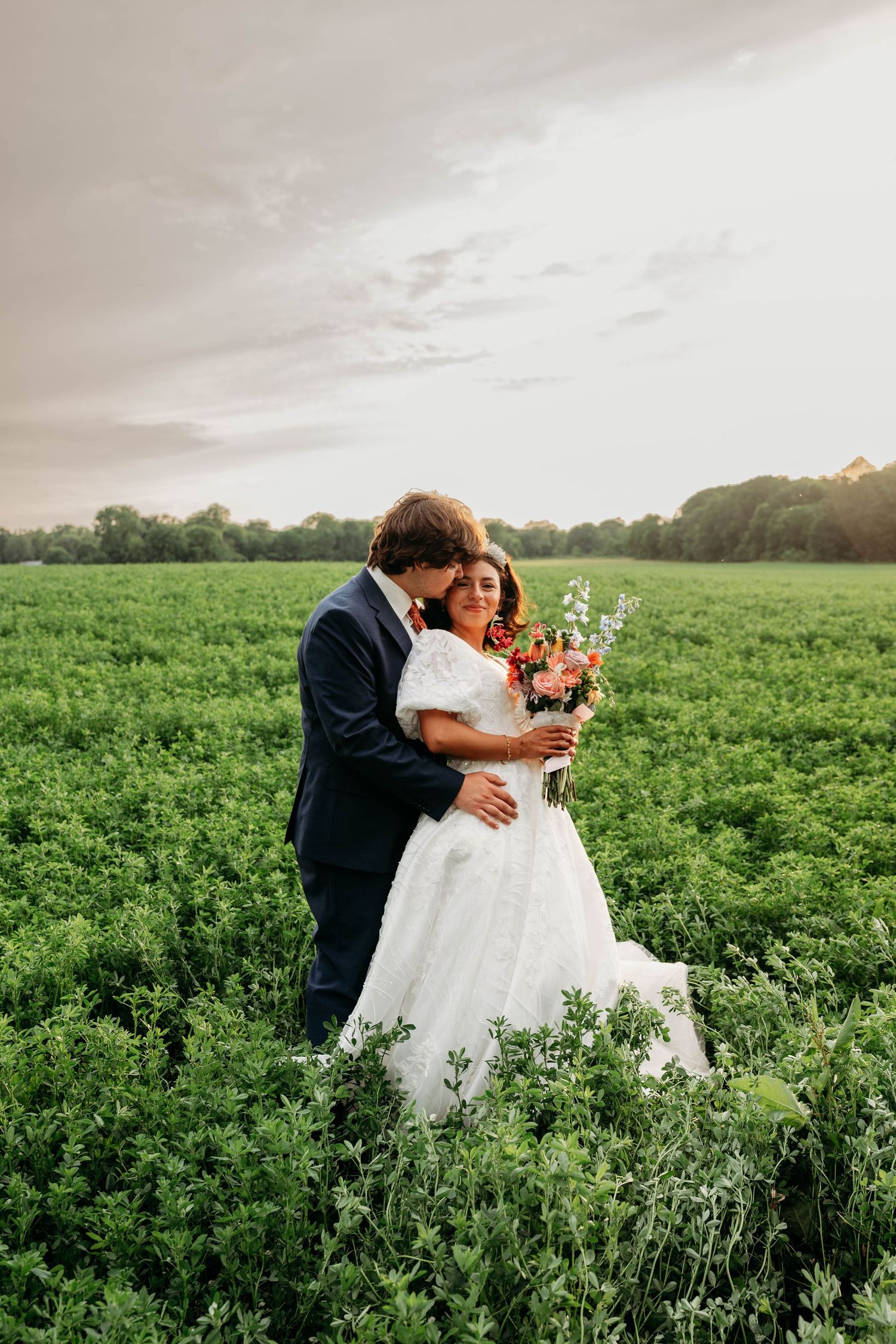 Bride and groom embracing in a lush green field, bride holding colorful bouquet, wearing white gown, groom in navy suit.