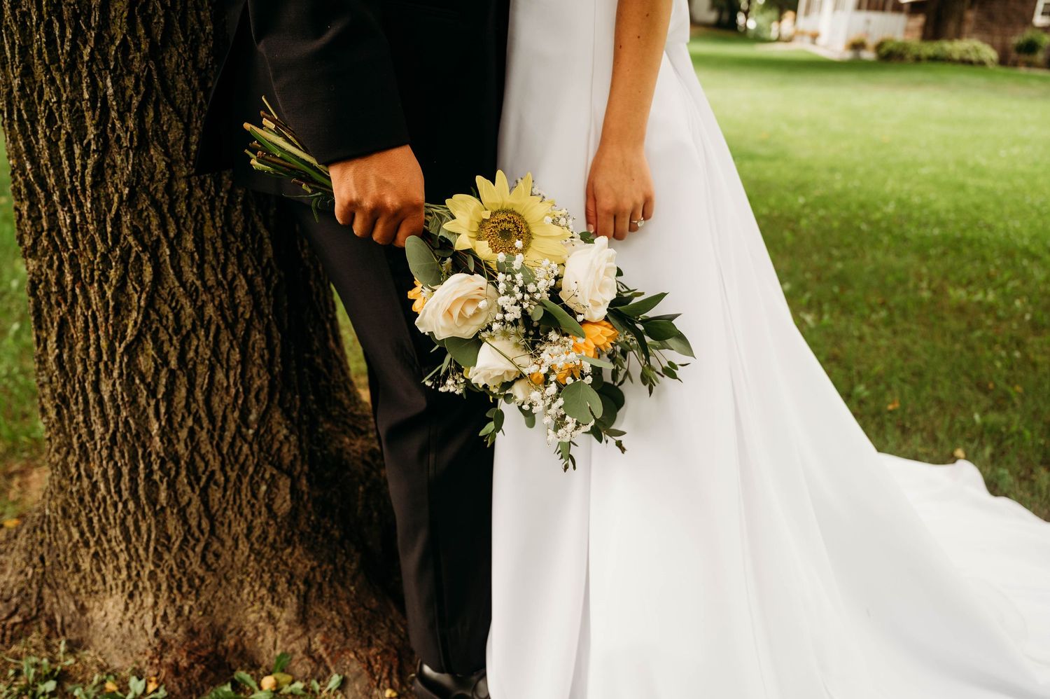 Bride and groom holding a sunflower wedding bouquet beside a tree on their wedding day.