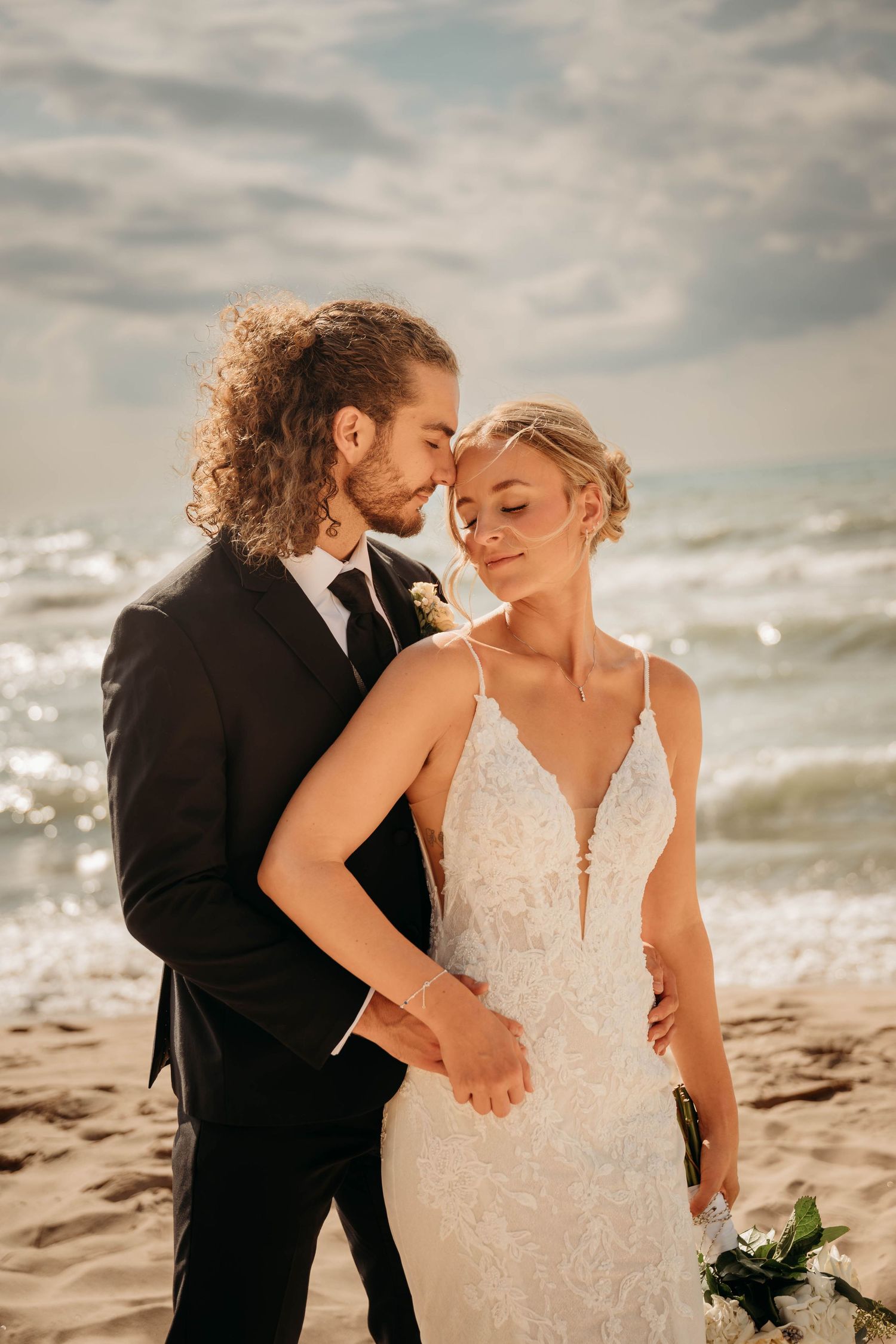 A bride and groom share a romantic moment on a sandy beach, with ocean waves and cloudy sky behind them.