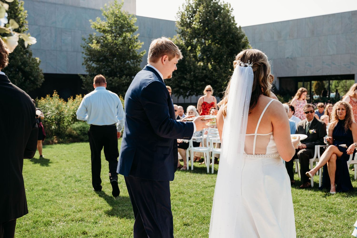 Groom in navy suit exchanges rings with bride in white gown during outdoor wedding ceremony on green lawn.