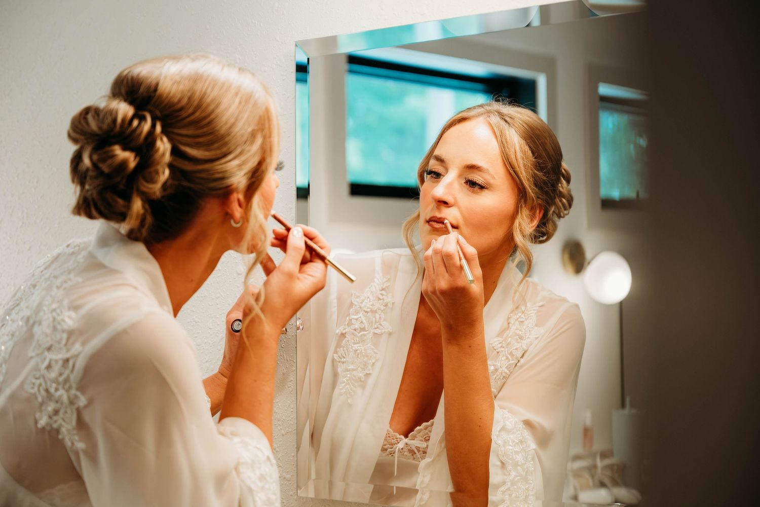 Bride in white lace robe applying lipstick while looking in a mirror during wedding day preparations.