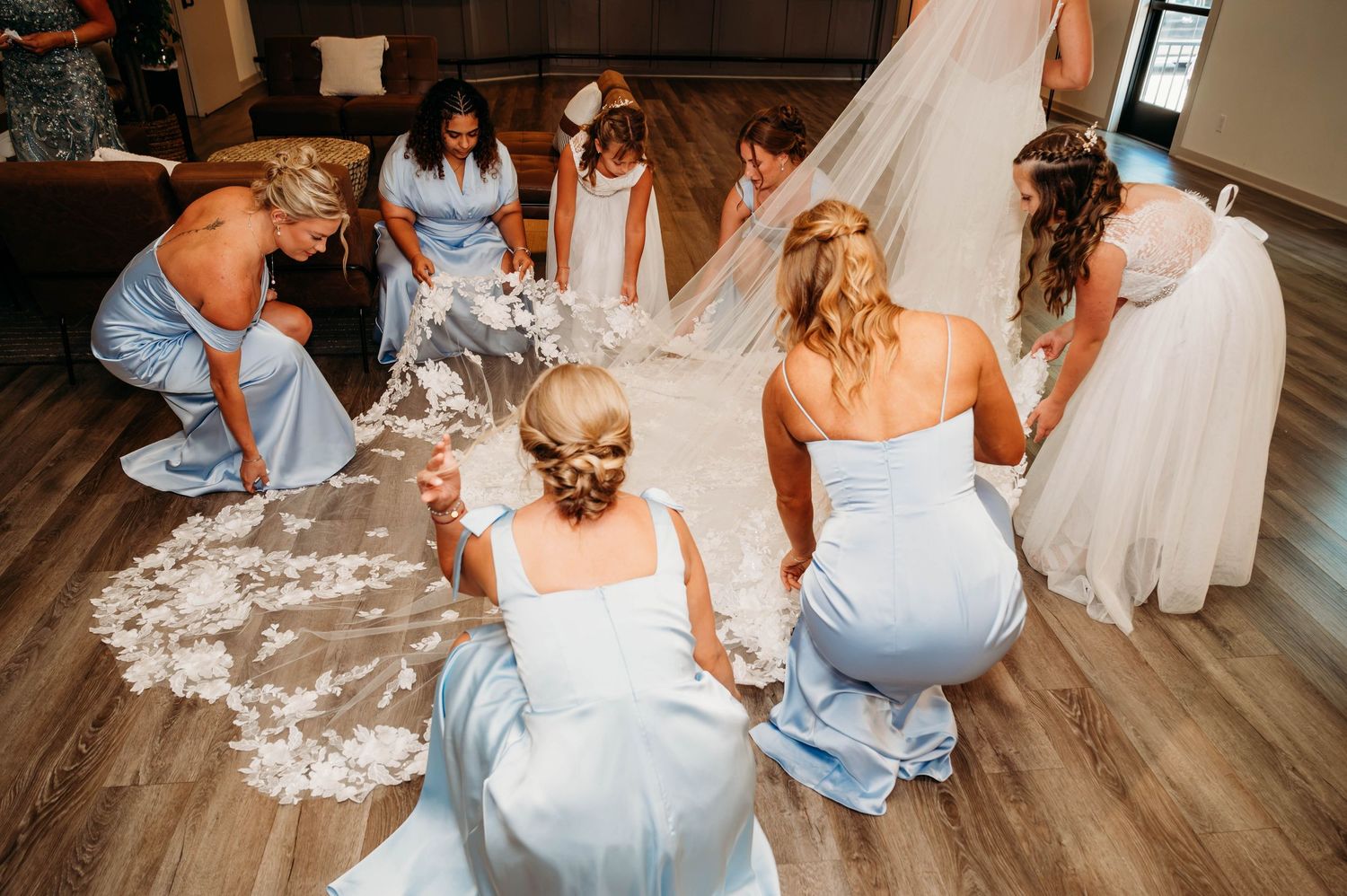 Bridesmaids in light blue dresses kneel to arrange a lace wedding veil and gown train on hardwood floor.