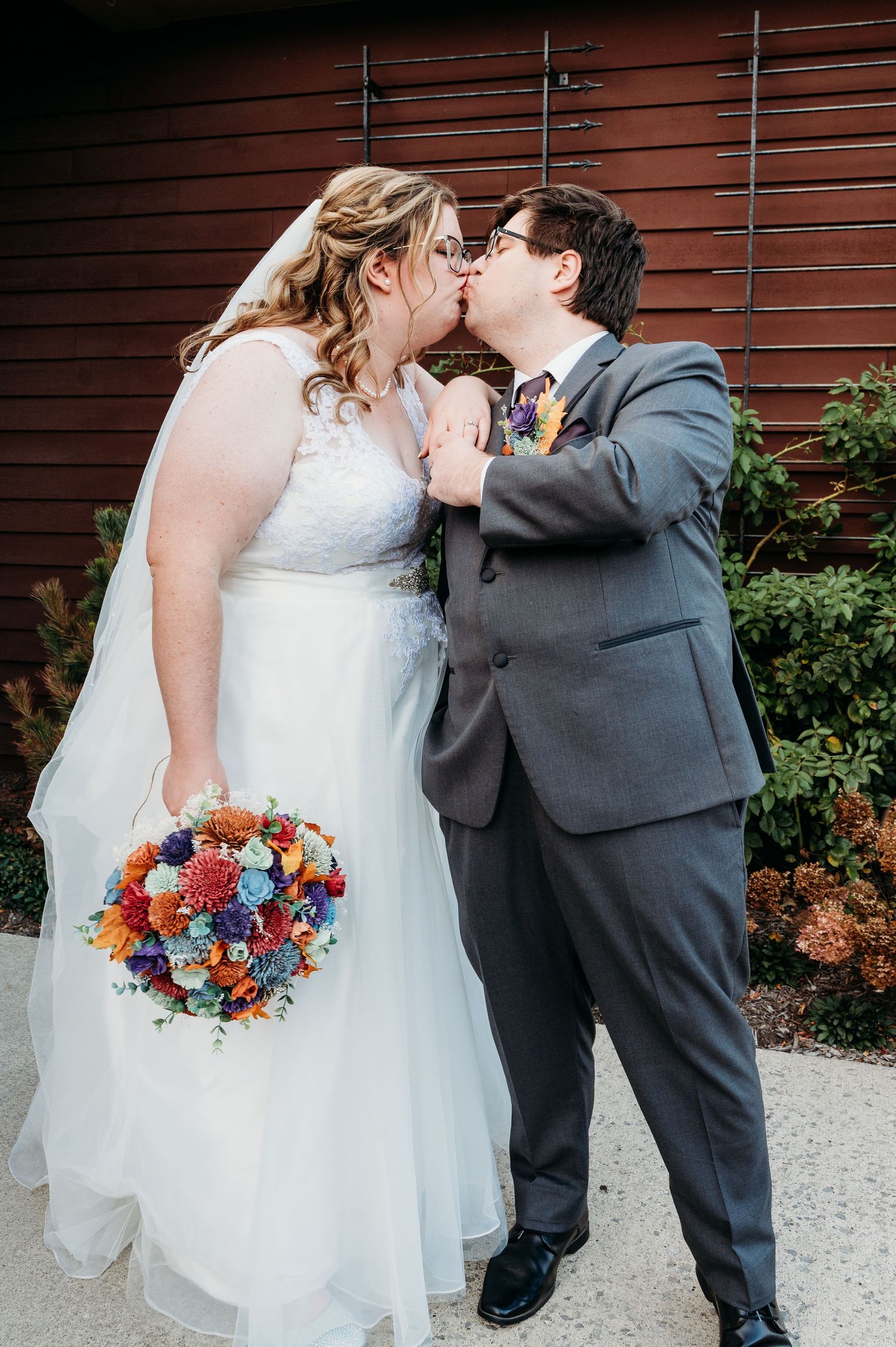 A couple in wedding attire share a kiss while holding a vibrant blue and orange bouquet against a red wall.