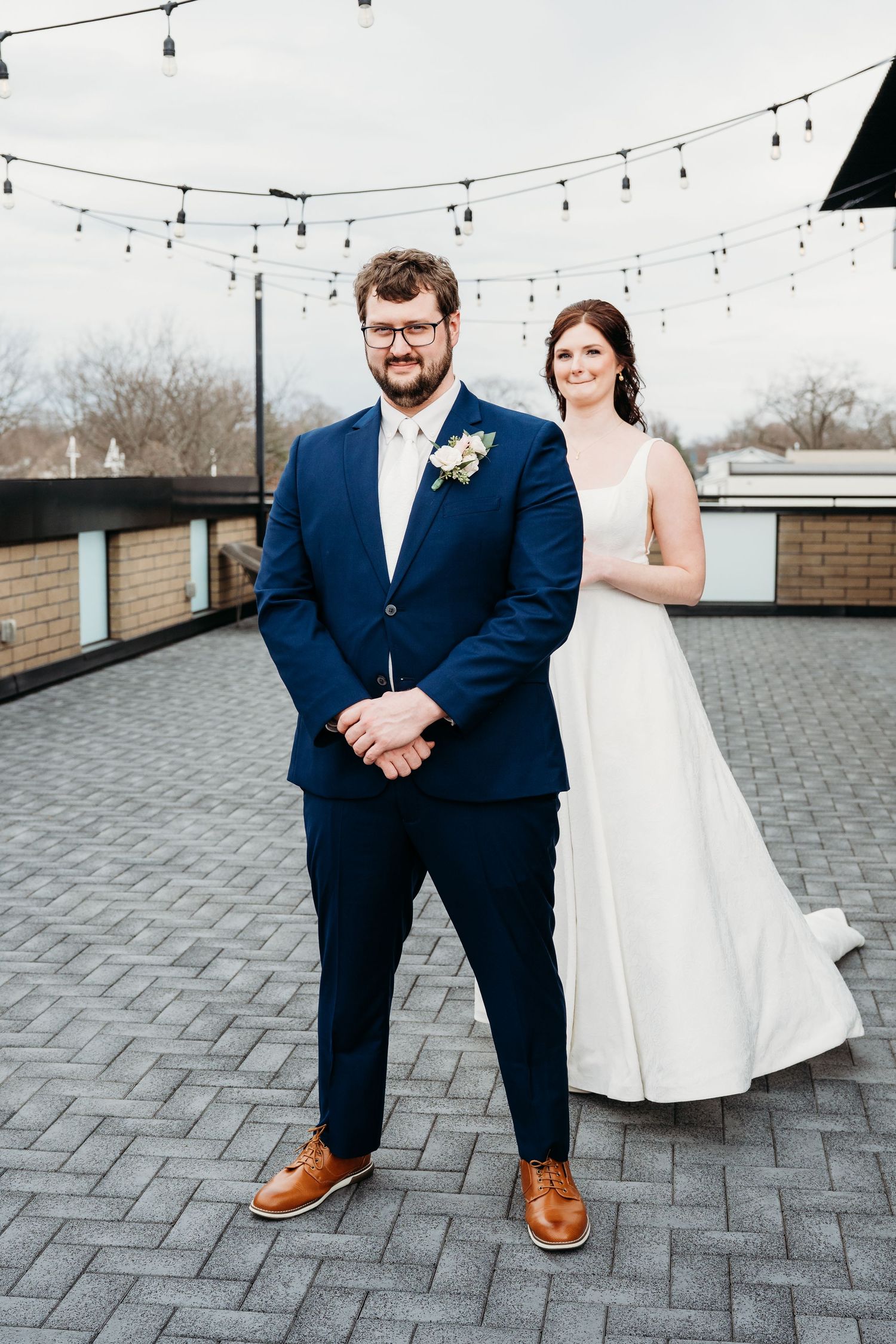Wedding couple poses outdoors on rooftop patio with string lights and brick walls in the background.