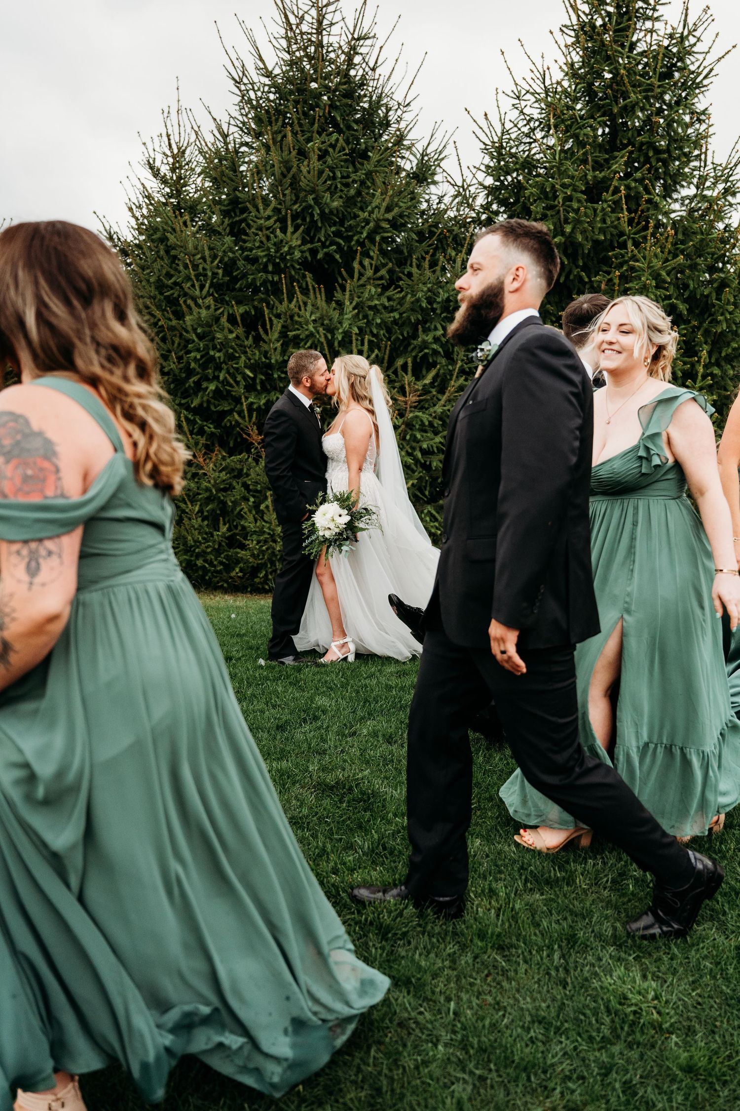 Wedding party walking across lawn with bridesmaids in sage green dresses and groomsmen in black suits.