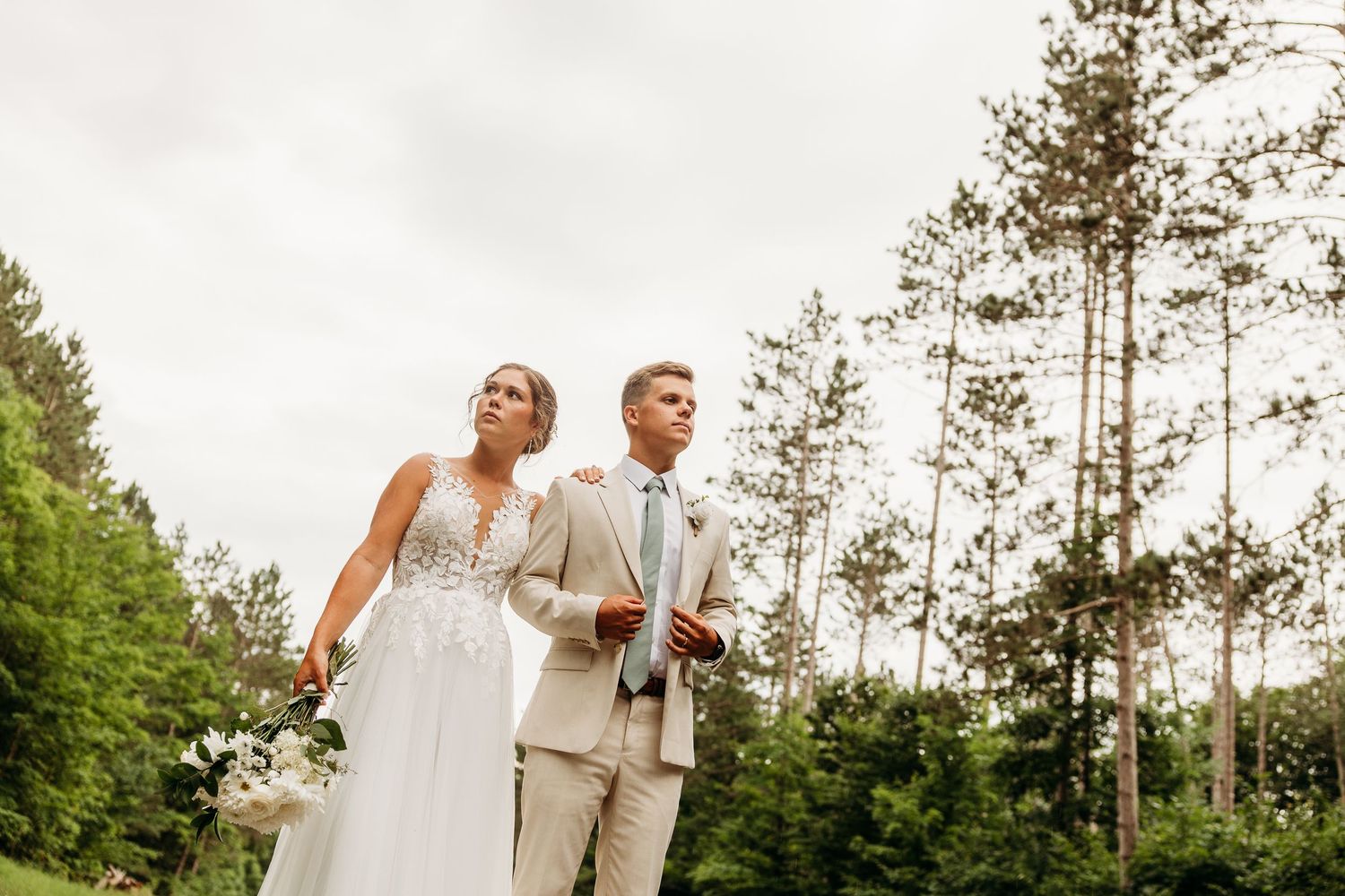 A couple in wedding attire stands together outdoors with tall pine trees and greenery in the background.