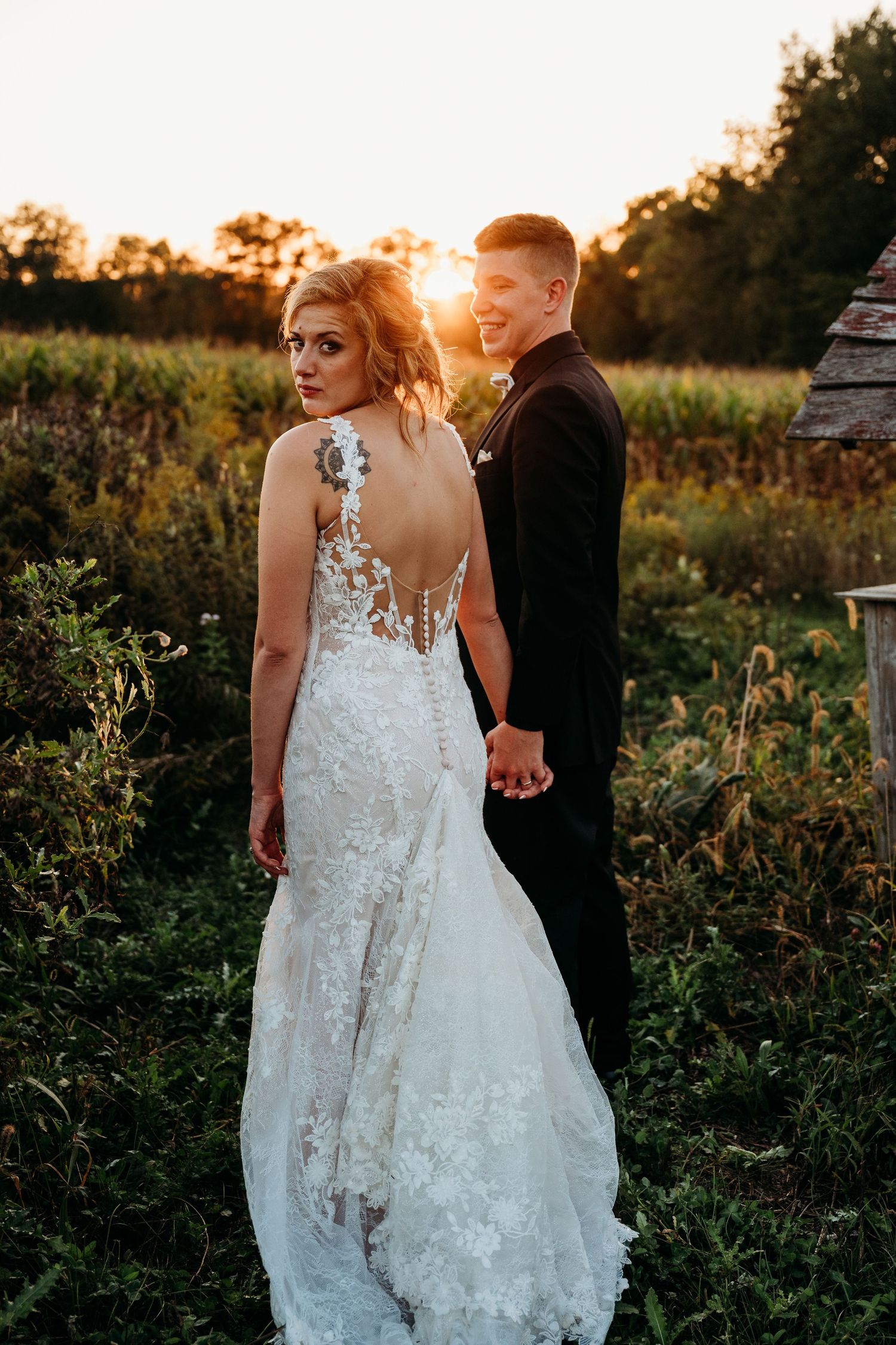 Bride in lace backless gown holds groom's hand at golden hour sunset in a lush outdoor field setting.