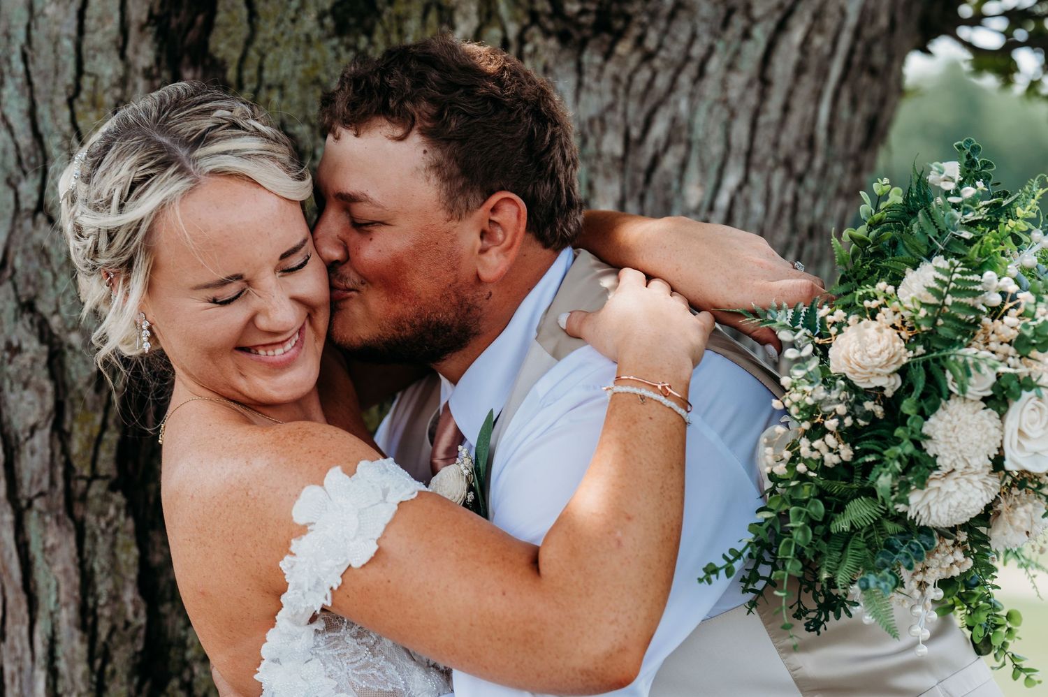 Bride smiling joyfully as groom kisses her cheek outdoors, holding a white and green wedding bouquet near a large tree.