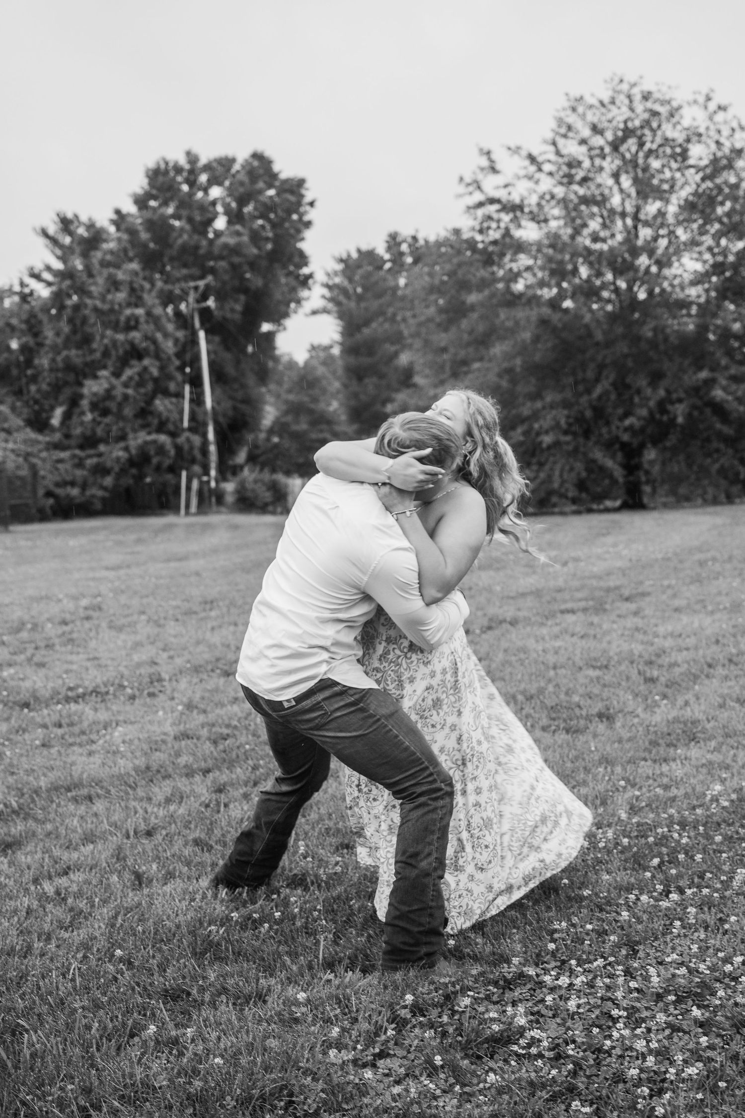 Black and white photo of couple embracing playfully in a grassy field outdoors.