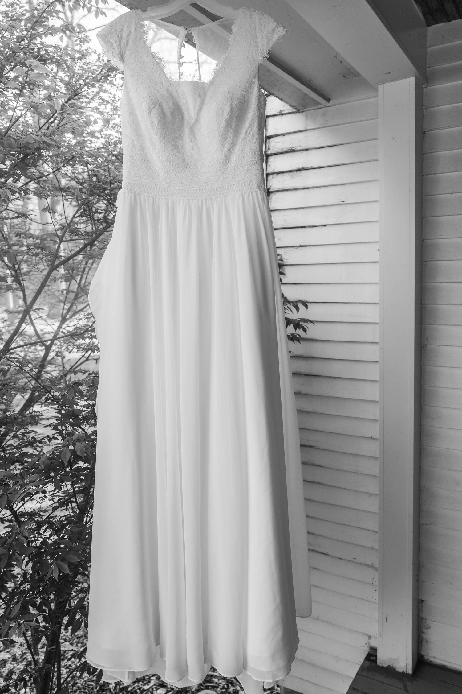Black and white photo of an elegant lace wedding dress hanging on a porch with shutters and trees in the background.