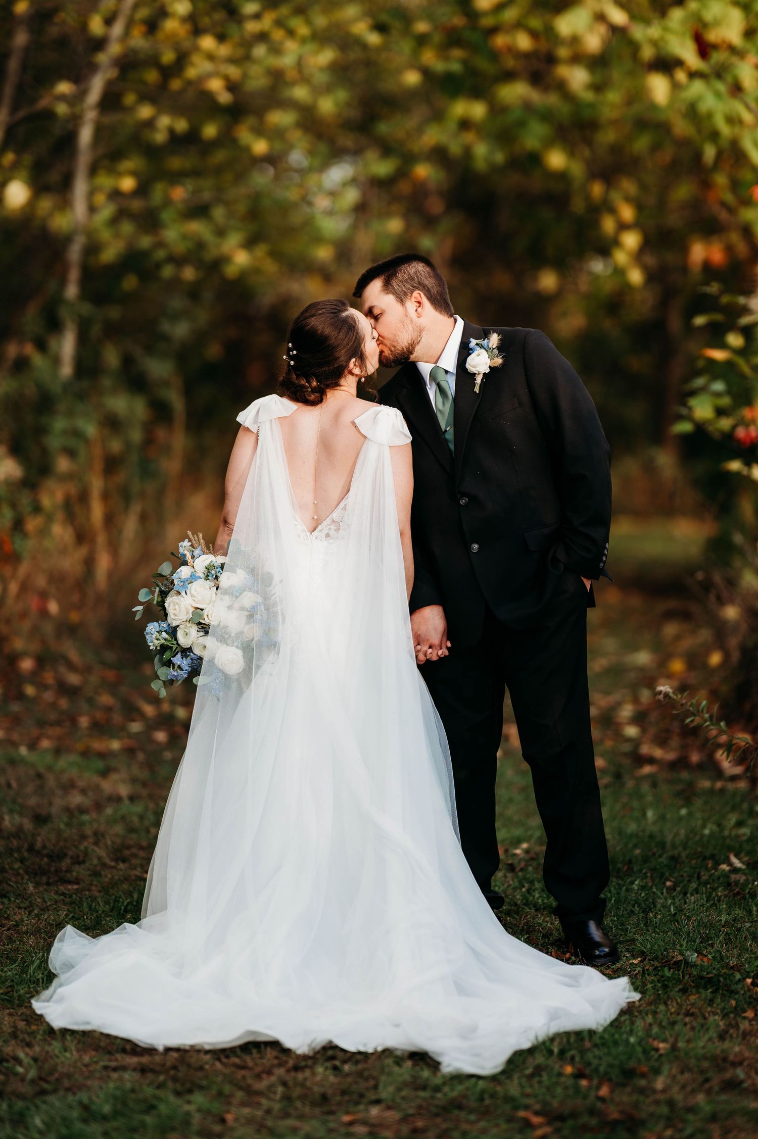 A romantic wedding couple shares an intimate moment in a wooded setting with autumn foliage in the background.