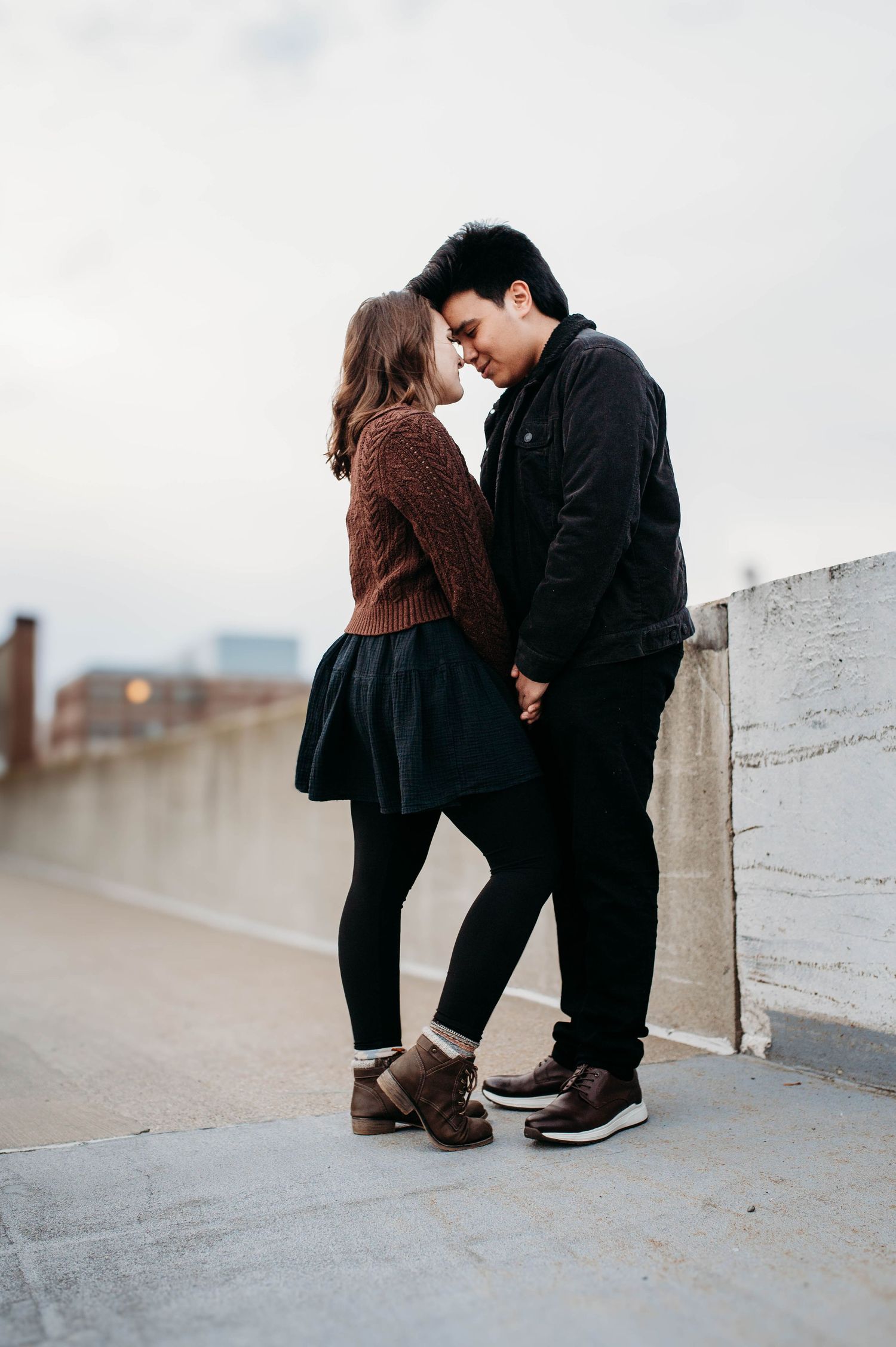 A couple in casual winter outfits share an intimate moment on a rooftop against an overcast sky.
