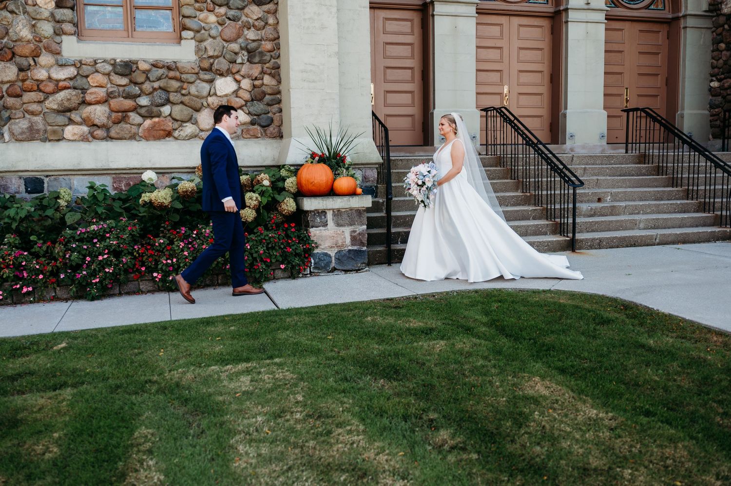 A couple in wedding attire poses outside a stone church with autumn decor and pumpkins on the steps.