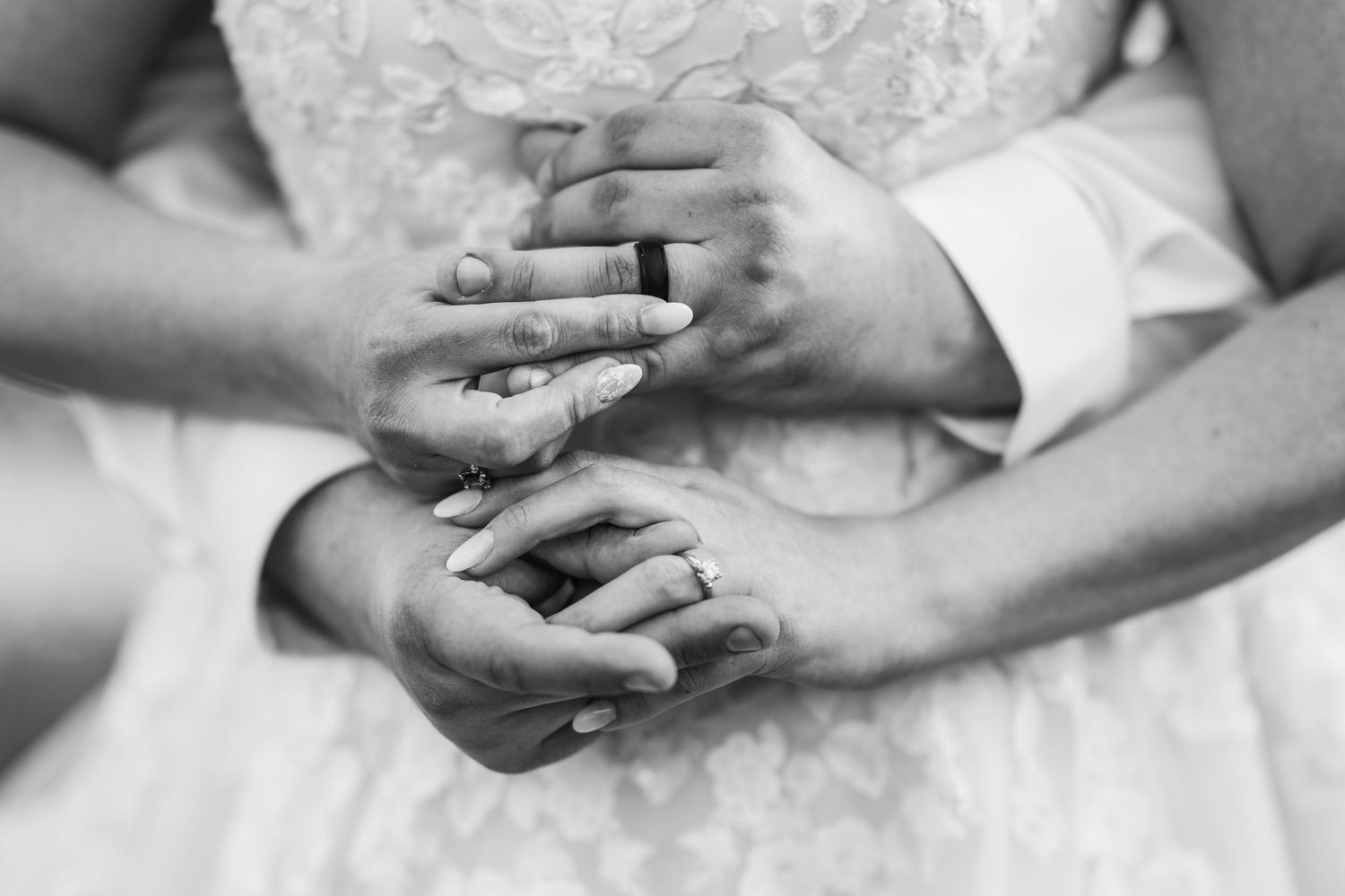 Black and white close-up of hands holding each other tenderly against a lace wedding dress background.