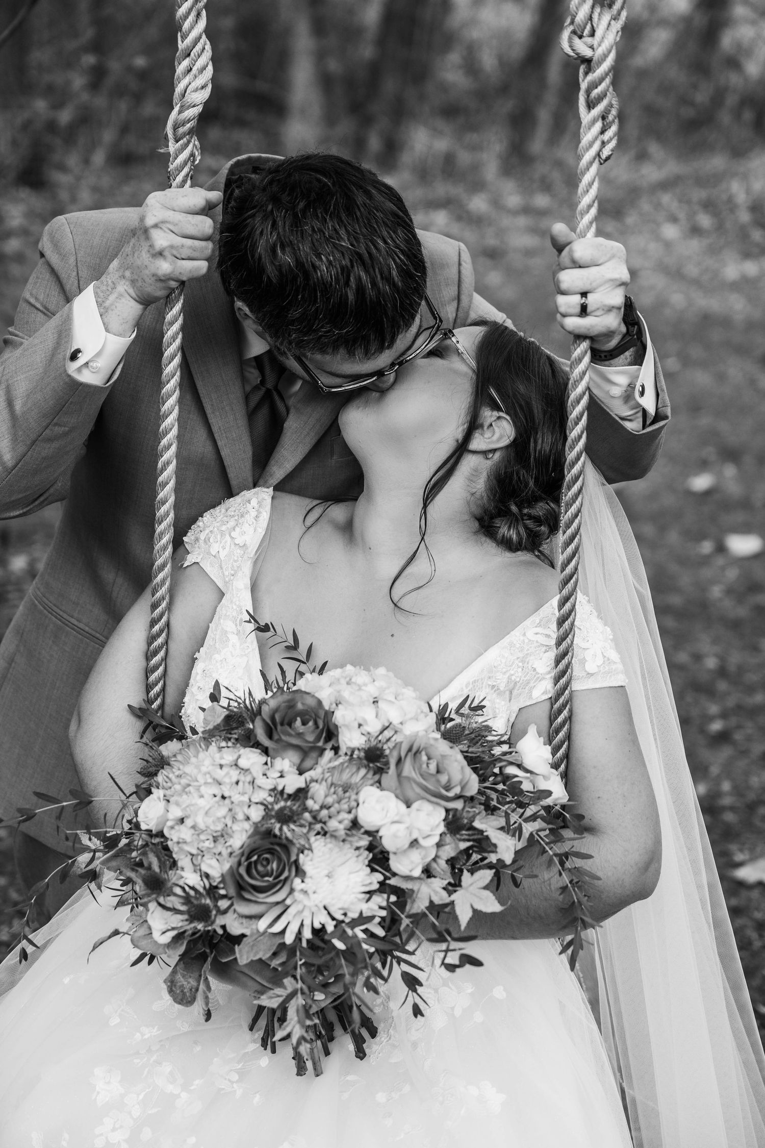 A romantic black and white wedding photo series showing a couple embracing on a swing with a beautiful floral bouquet.