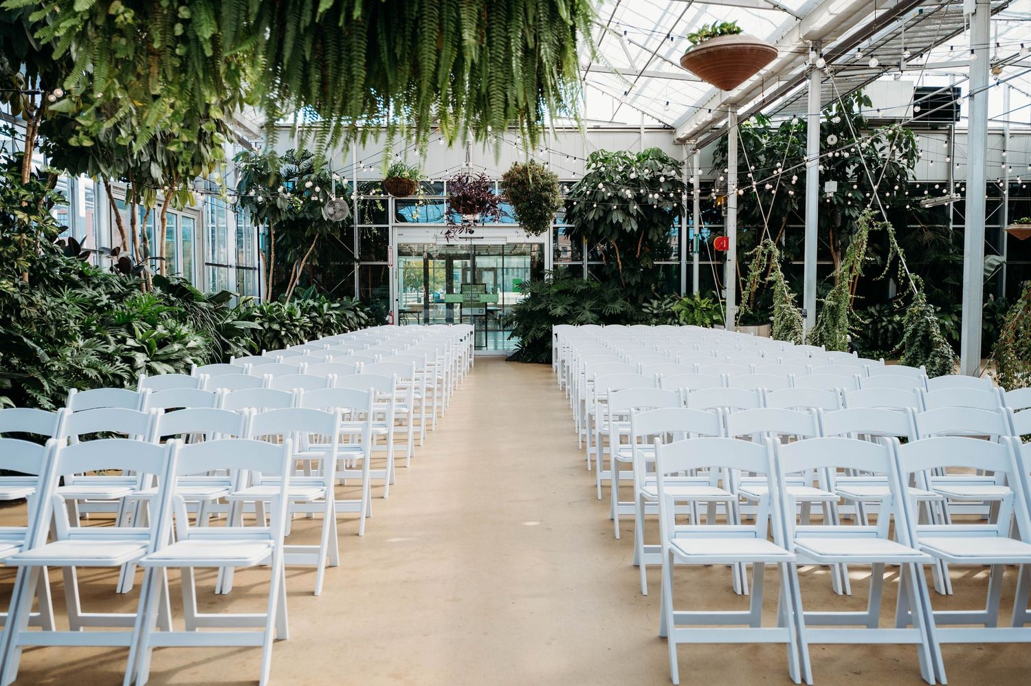 White folding chairs line an aisle in a greenhouse wedding venue with hanging plants and natural lighting.