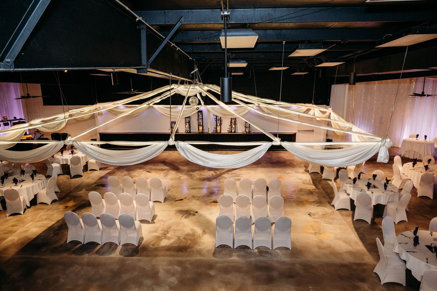 Wedding ceremony venue setup with white chairs arranged in rows under a draped fabric ceiling with elegant lighting.